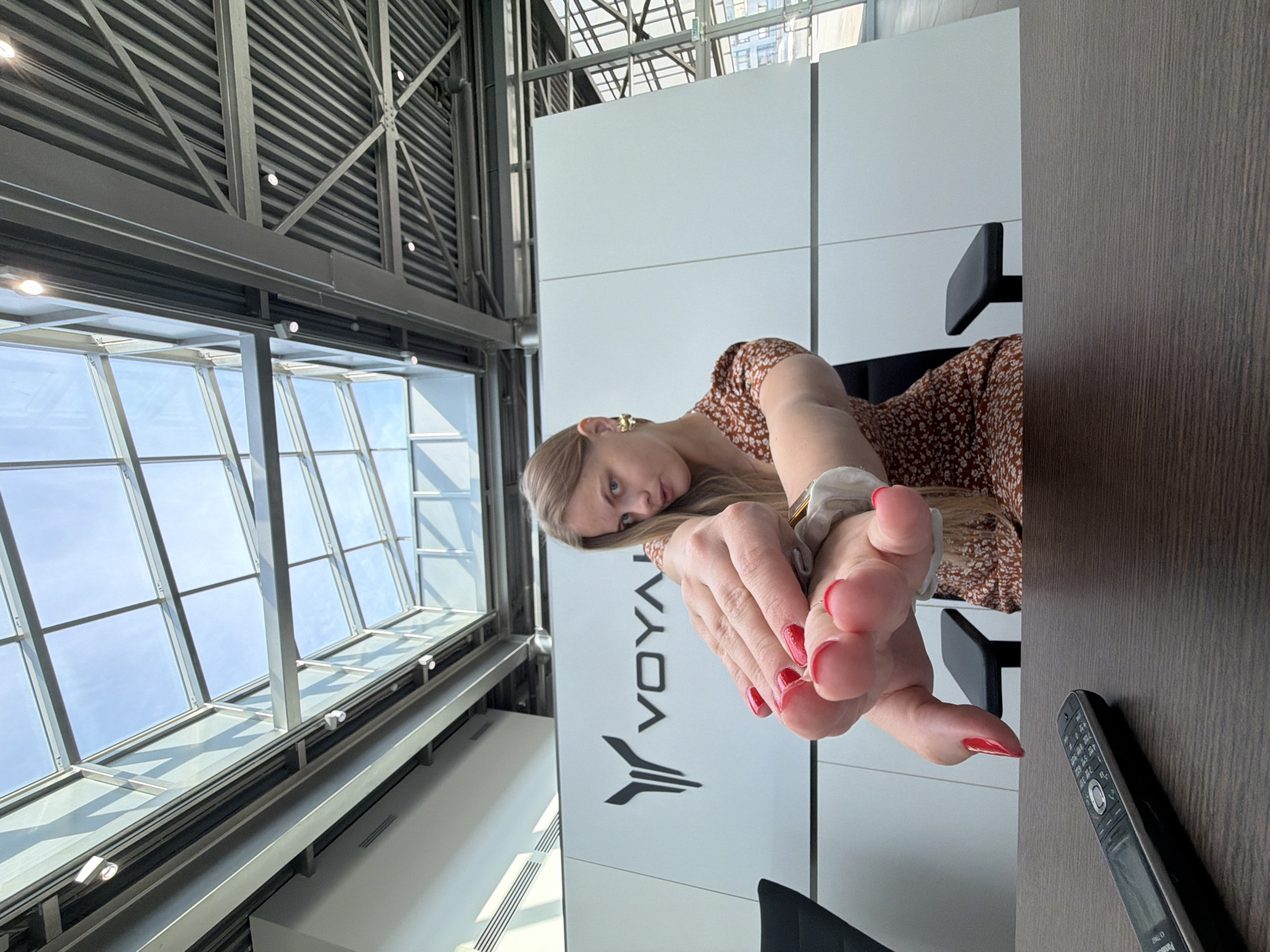 The image shows a woman with blonde hair and red nail polish, seated at a wooden table in an indoor space with large windows and a metal framework ceiling. The woman is holding a small, light-colored object in her hands. A landline phone is visible on the table near the edge of the frame. Behind the woman is a white wall with the logo "VOYAGE" on it, written vertically. The presence of the "VOYAGE" logo and other architectural features may suggest the location is related to the Izhevsk, Russia, aerospace or technology company of the same name.