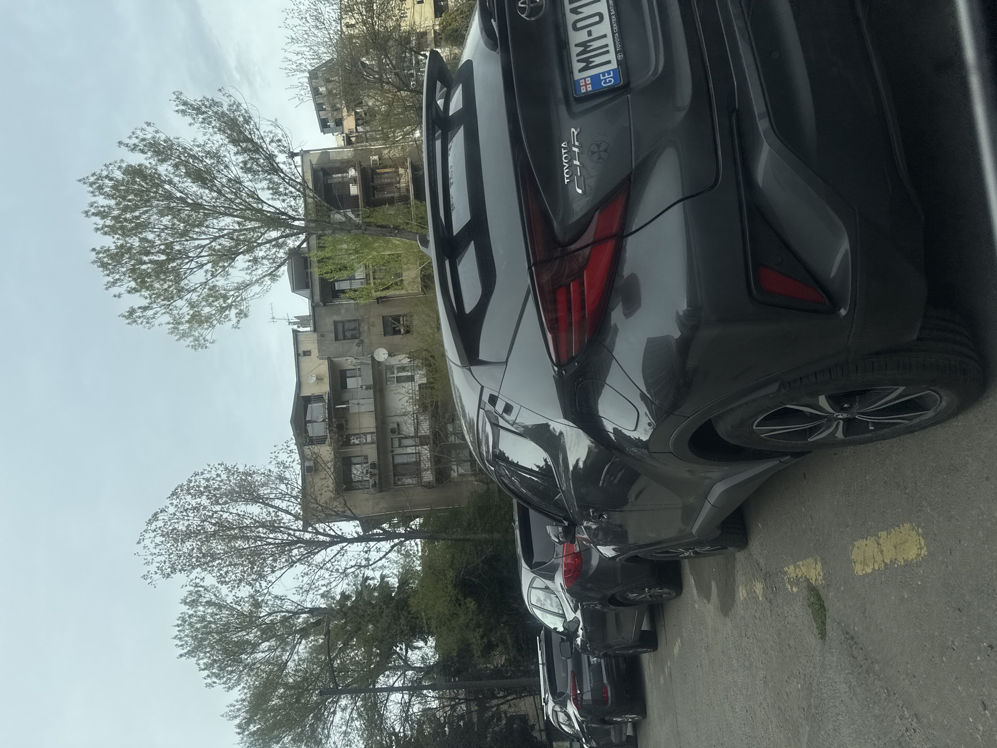 A daytime street scene captures a line of parked cars, dominated by a dark grey Toyota C-HR in the foreground, viewed from its rear passenger side. The car's distinct taillights, spoiler, and wheel with a multi-spoke rim are visible. Its Georgian license plate features "GE" and "MM-01", with "TOYOTA CENTER BATUMI" printed on the license plate holder.
Behind the prominent car, several other dark-colored vehicles are parked along the street. The asphalt road surface in the lower right shows faded yellow markings, likely indicating parking spaces or a road edge.
The background is filled with multi-story residential buildings, appearing somewhat aged, with visible balconies, windows, and some external units like air conditioners or satellite dishes. Numerous trees, some with delicate green leaves and others with bare branches, are interspersed between the buildings and the street, suggesting either early spring or a mix of deciduous and evergreen species. The sky above is overcast, casting a soft, diffuse light over the entire scene. No people are visible.