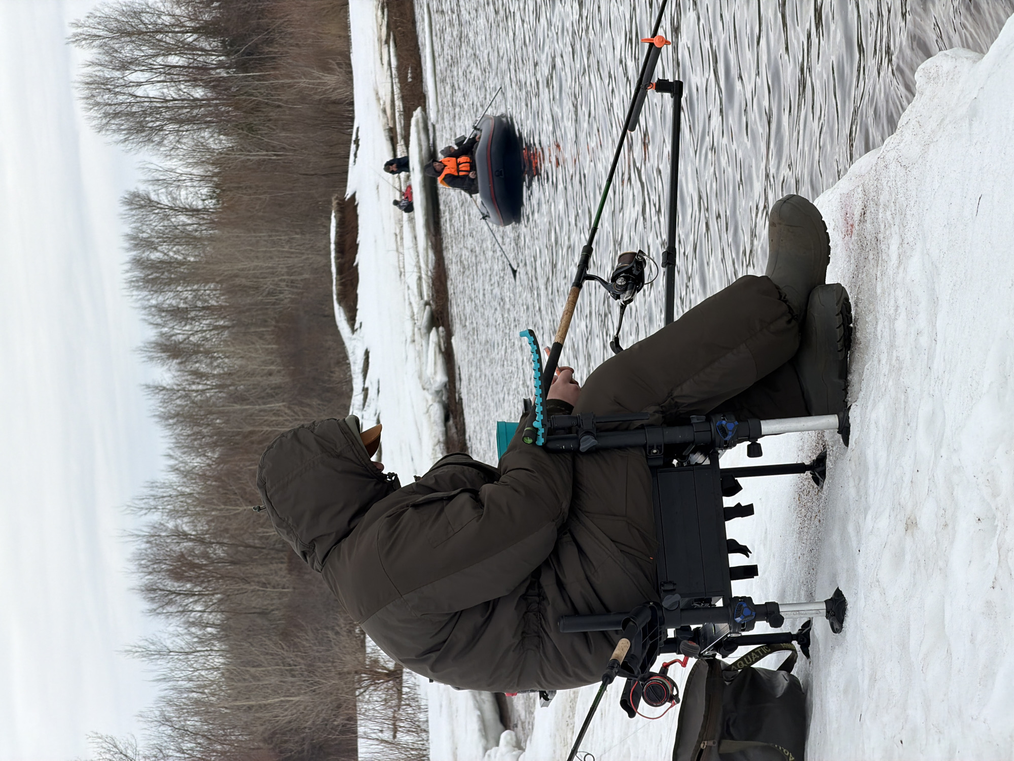 The image depicts a person ice fishing, likely in the city of Chaikovsky, Russia, given the context.
**People:** One person is shown seated and engaged in fishing. Two other people are visible in an inflatable raft further out on the body of water. One of the people in the raft is wearing a bright orange life vest.
**Subjects:** The primary subject is the seated person fishing through a hole in the ice. The secondary subject is the people in the raft.
**Objects:** Notable objects include fishing rods, reels, and equipment, a fishing platform seat, a raft, ice, snow, and surrounding vegetation. The seated person is wearing waterproof clothing and boots.
**Actions:** The seated person is actively fishing. The people in the raft appear to be traveling on the water.
**Interactions:** There is an implied interaction between the people fishing, as they are sharing the same body of water.
**Environment:** The environment is snowy and cold, indicating winter. The body of water is partially frozen over with ice, with open sections of water. The trees on the shoreline are bare, further indicating the winter season.