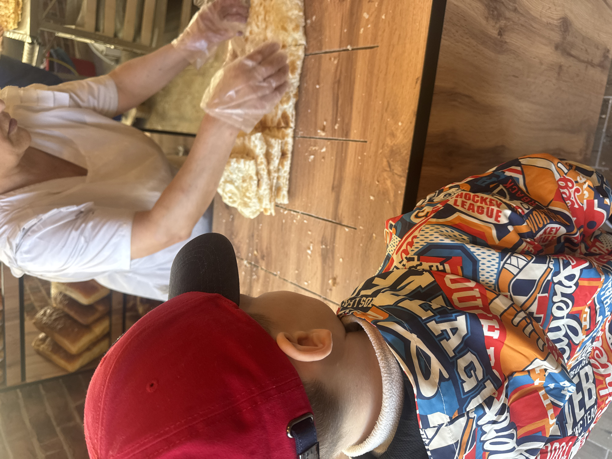 An overhead, slightly angled shot depicts a food preparation scene, likely within a bakery or market stall, featuring two individuals. On the left, a person wearing a white shirt and clear plastic gloves is actively engaged in handling a long, golden-brown piece of flatbread, possibly lavash. Their hands are positioned over the bread, which rests on a rich, warm-toned wooden counter. Parallel score marks or cuts are visible on the wooden surface, suggesting the bread has been, or is being, sectioned.
On the right side of the frame, another individual, seen from behind and the side, observes the activity. This person wears a red baseball cap, turned backward, and a brightly colored, patterned jacket. The jacket features a busy design with various stylized letters and words, including visible fragments like "HOCKEY LEAGUE," "BACK," "WORLD," and "KC TEAM," indicating a sports or team-related theme. The person's ear is visible between the cap and the jacket's collar as they lean forward, looking towards the flatbread on the counter.
In the background, behind the person in white, several loaves of baked bread are stacked on a metal rack, reinforcing the setting as a place that sells freshly made baked goods. The scene appears to be an indoor setting, likely in a food establishment in Sochi, Russia, with a focus on the interaction between the food preparer and the observer.