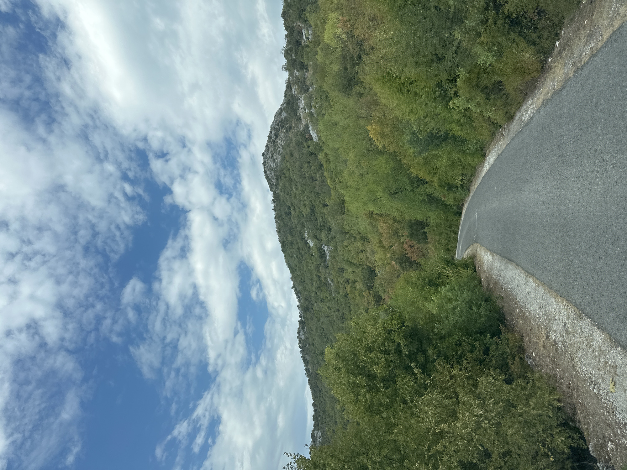 The image depicts a winding asphalt road leading through a verdant landscape with a mountain range in the background. The sky above is a bright blue, dotted with scattered white clouds. Dense green foliage lines both sides of the road, with the trees growing up the slopes of the mountain. The mountain itself is covered in similar vegetation, with rocky outcrops visible at its peak. The road appears to be in a rural or mountainous area, with no visible signs of human habitation or activity other than the road itself. The scene suggests a natural and relatively untouched environment, likely in a region with a hilly or mountainous terrain. The location is stated to be Kruševica, Montenegro, indicating a Balkan setting.