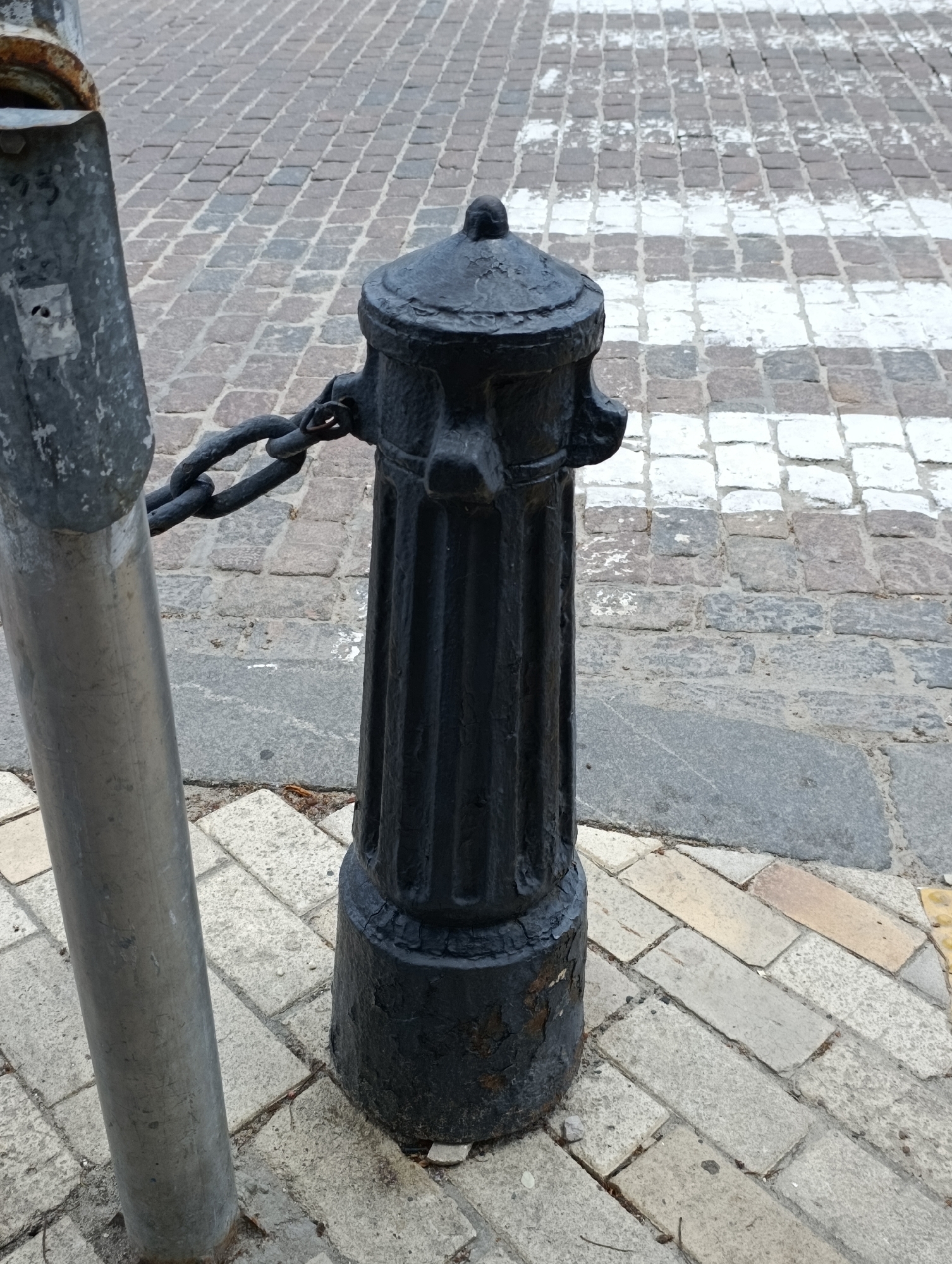 This is a medium shot, eye-level, outdoor photograph of a black cast-iron bollard with a chain attached to a metal post. The bollard is located on a pedestrian crossing with cobblestone paving. The bollard is fluted and has a cap-like top. A thick, dark chain connects the bollard to a silver metal post on the left side of the frame. The cobblestones are of various sizes and colors, ranging from gray to brown and white. White stripes, indicative of a pedestrian crossing, are visible on the cobblestones behind the bollard. The image appears to have been taken in a city environment, possibly a public square or street. The lighting suggests an overcast day. There are no people or animals visible in the image. The general condition of the bollard and post suggests wear and tear, with some rust visible on the metal post and chipping paint on the bollard.