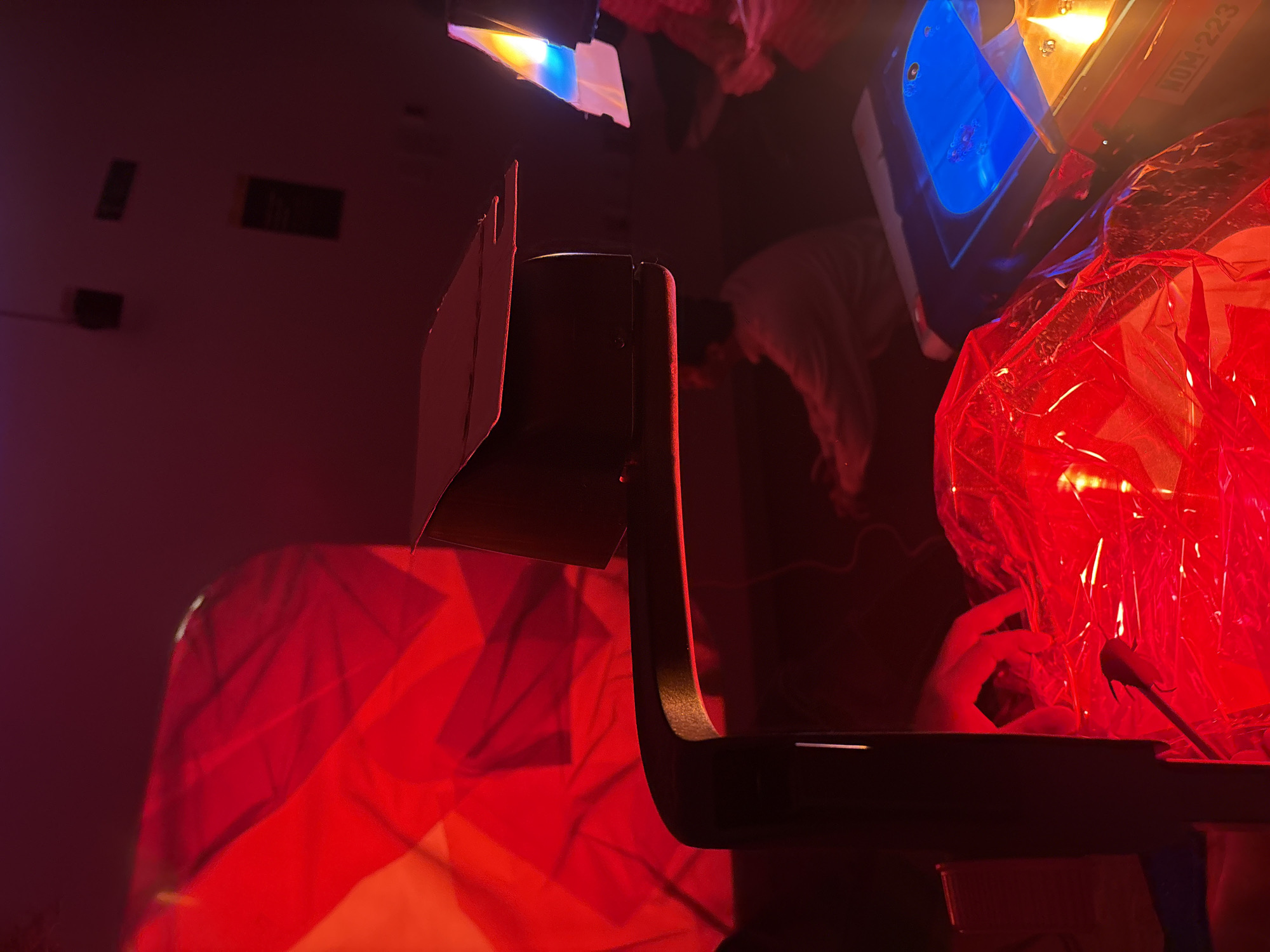 The scene depicts an indoor environment illuminated predominantly by artificial red light, characteristic of an artistic installation, experimental setup, or darkroom. The location is identified as Mexico City, Mexico, based on the partial "NOM-223" label visible on equipment, referencing "Normas Oficiales Mexicanas" (Official Mexican Standards).
In the immediate foreground, to the left, a dark, curved, possibly metallic support arm or structure is present, exhibiting a subtle red glow along its lower edge. Beneath this structure, a transparent, crinkled red plastic material is brightly illuminated, with a human hand visible, reaching into or manipulating this material from the left side of the frame.
Midground center features a dark, rectangular overhead projector or similar display device, with its top flap partially open, casting a dim light.
In the background, to the left, a large, angular, red geometric pattern is projected onto a dark wall or screen. To the center-right, a blurred figure of a person, likely an adult, is seated or crouched, facing away from the viewer, wearing a light-colored long-sleeved garment. They appear to be interacting with unseen equipment below them. Adjacent to this person, on the far right, another light source emits distinct blue and yellow-orange light. Below this light source, a piece of dark equipment displays the partial white alphanumeric label "NOM-223". The overall environment is dark, emphasizing the various colored light sources and projections.