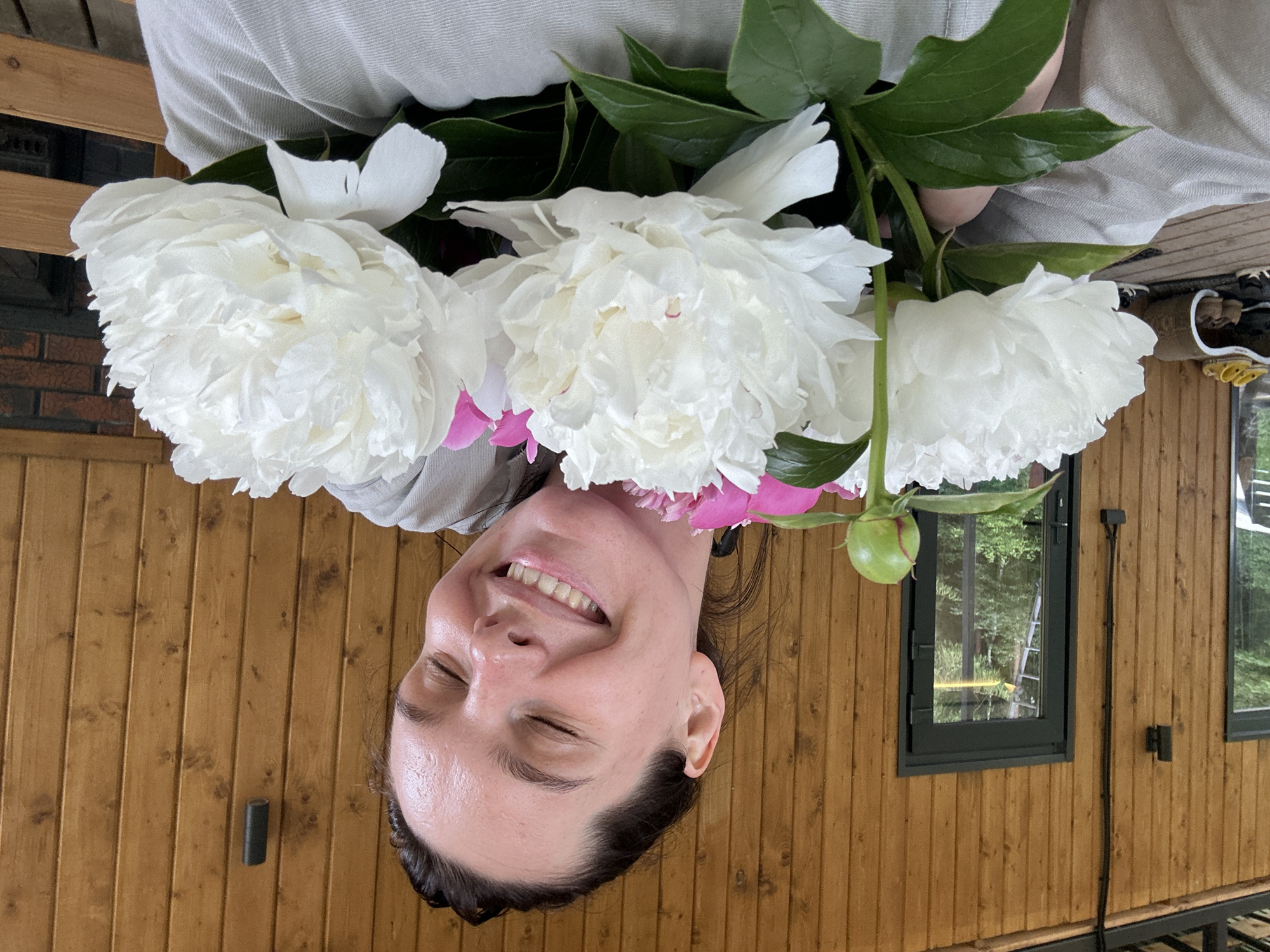 A woman is holding a bouquet of white and pink peonies in front of a wooden wall. She has her eyes closed and is smiling. The scene appears to be outdoors, on a porch or deck, with a wooden structure and some greenery visible in the background. The overall impression is one of happiness and relaxation. The location is not specified in the image or the provided crops.