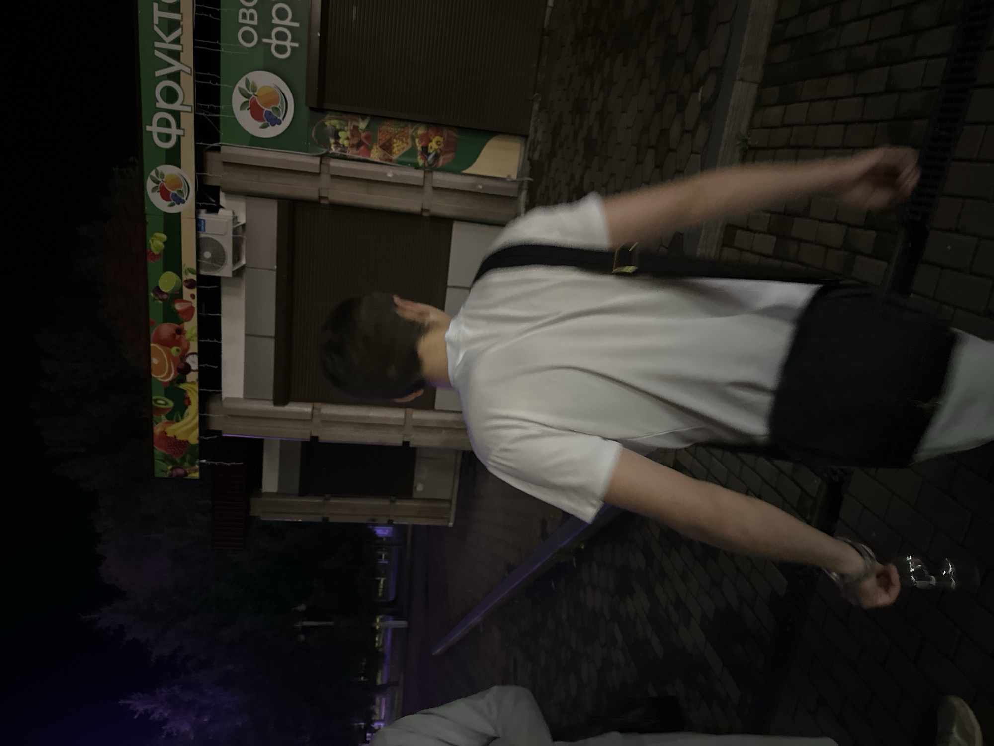 The image depicts a person from behind, wearing a white t-shirt and a black strap across their chest. They are standing on a paved walkway, which appears wet, suggesting it might have recently rained or is damp. Behind them, there's a building with closed shutters and a storefront adorned with colorful images of fruits, including oranges, apples, and grapes. The signage above the storefront has Cyrillic text, with "ФРУКТЫ" (FRUITS) clearly visible. The setting appears to be a street or market area at night, indicated by the darkness and artificial lighting. The person's arm is extended forward, and their hand is blurred, suggesting movement. There's another person partially visible in the foreground, wearing a light-colored garment. The overall mood is ambiguous due to the low light and the blurred action.