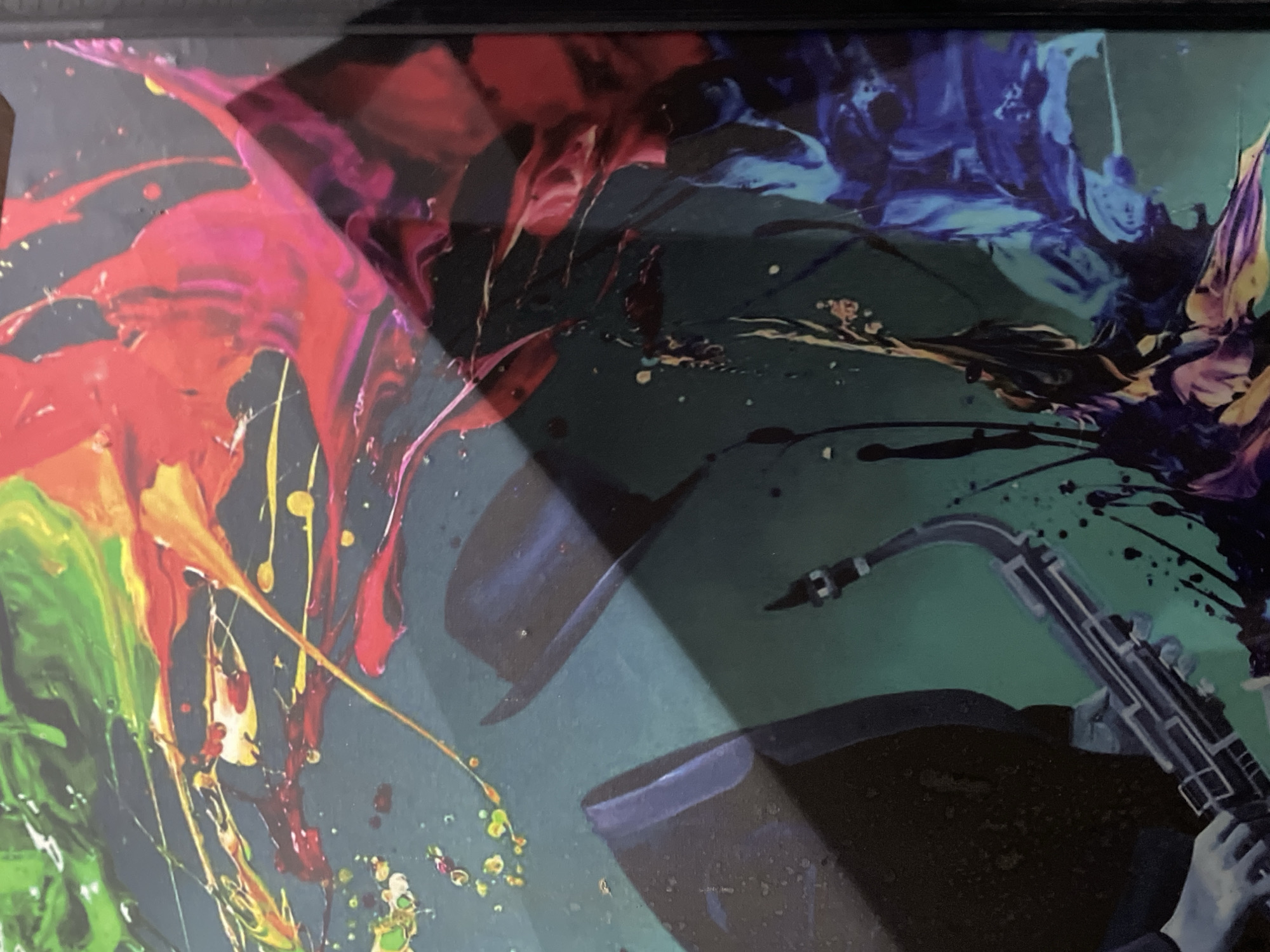 The image is a close-up of a framed abstract painting. The left side features vibrant colors, including green, red, and yellow, appearing as fluid splatters. The right side displays darker hues of blue and purple, also resembling fluid forms. In the bottom right corner, a saxophone is partially visible, along with what appears to be a hand holding it. There are also abstract shapes that could represent a person wearing a hat. The background color is a muted green.
There is no information in the image to suggest it is located in Abuja, Nigeria, or any other specific location. The image focuses solely on the artwork.