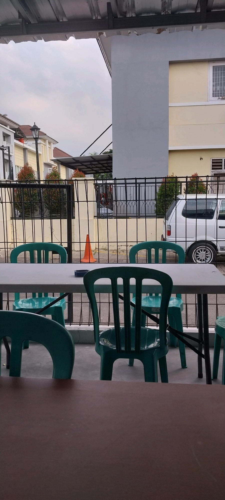 The image depicts an outdoor seating area with several green plastic chairs arranged around a light-colored table. Behind the table, a black metal fence encloses a yard with a white van parked to the right. An orange traffic cone is visible near the fence. In the background, houses with yellow and white facades and red-tiled roofs are visible, along with a street lamp. The sky is overcast, suggesting a cloudy day. The overall scene appears to be a casual outdoor dining or gathering space, likely in a residential area of South Tangerang, Indonesia.
