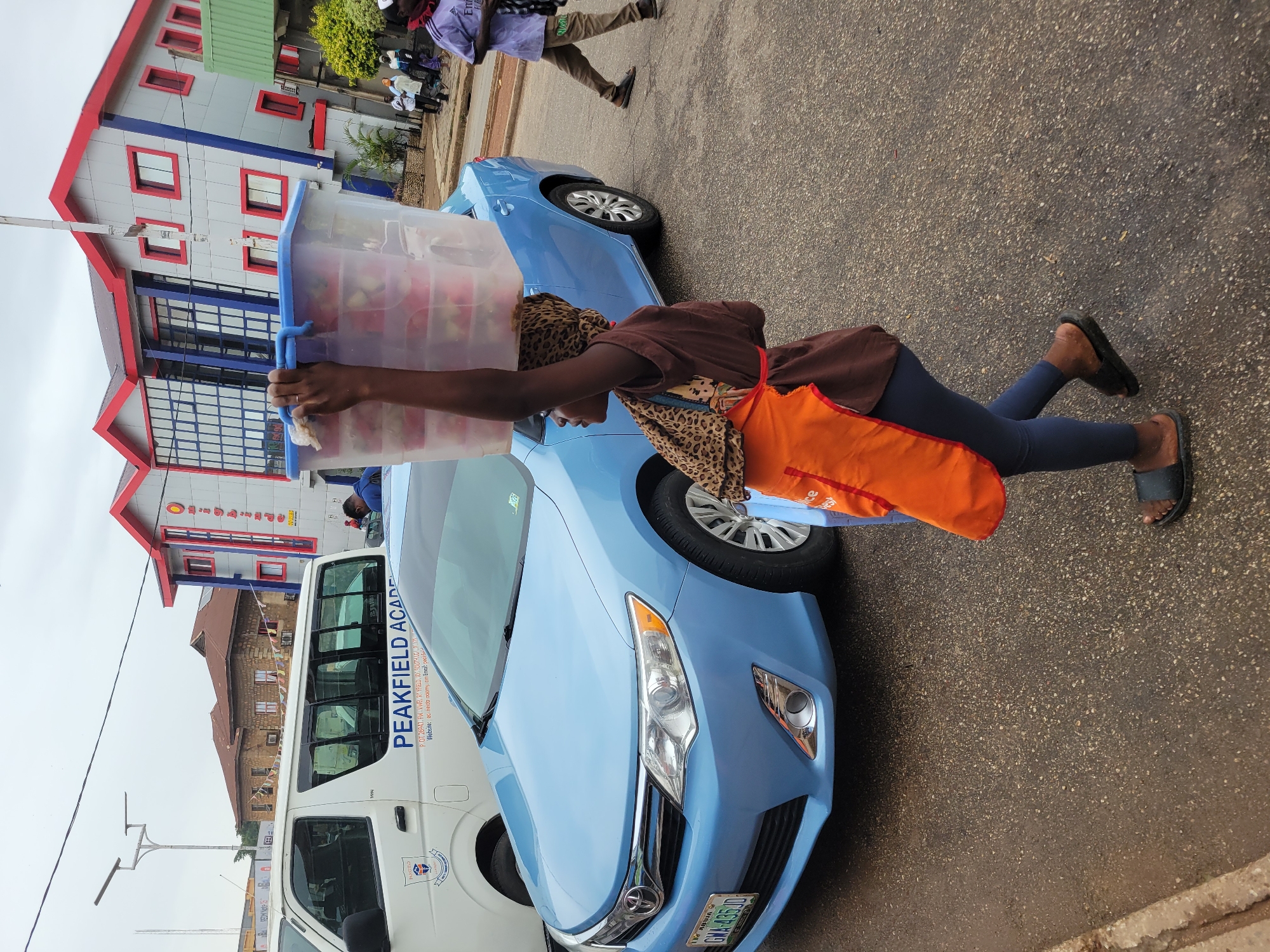 A woman wearing a leopard print headscarf, brown shirt, blue leggings, an orange apron, and flip-flops carries a large clear plastic container filled with what appears to be fruit. The container is positioned over a light blue sedan, which is parked next to a white van with "PEAKFIELD ACAD" printed on the side. A building with red and blue architectural details is visible in the background. The location appears to be in Jos, Nigeria, based on the license plate visible on the car. Another person is walking in the background.