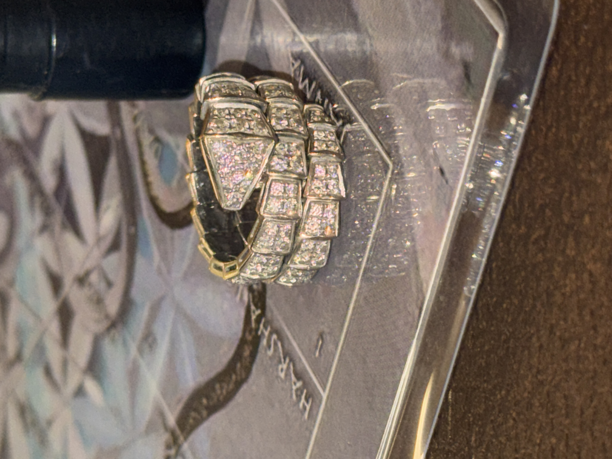 A close-up shot shows a gold and diamond snake-shaped ring lying on what appears to be a glossy magazine or book cover. The ring, crafted in a winding serpent design, features diamond-encrusted segments. The magazine or book cover depicts a snake drawing in a gray tone, with some letters visible. The scene takes place indoors. No specific details on the location of North Kuta, Indonesia are discernible from the image. There is also a black object at the very top of the image, it's a dark, possibly black, object that's partly cropped, with some cylindrical features and darker edges.