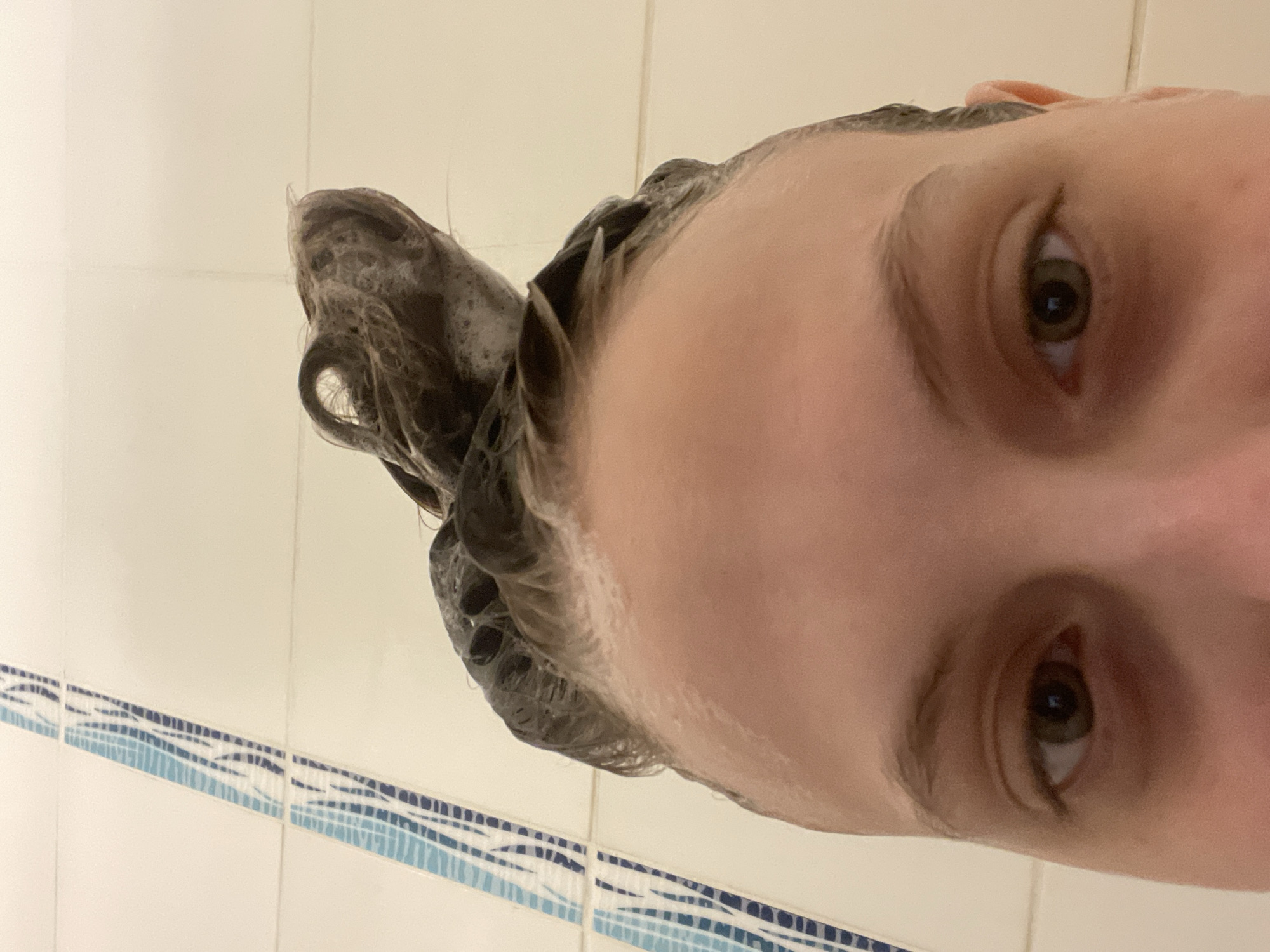 A close-up, vertically-oriented image shows a person's face with wet, dark hair piled on top of their head. The person is looking at the camera with their brown eyes. The background consists of white tiles with a blue decorative border, suggesting a bathroom setting. The image does not contain any information indicating its location in Ufa, Russia.