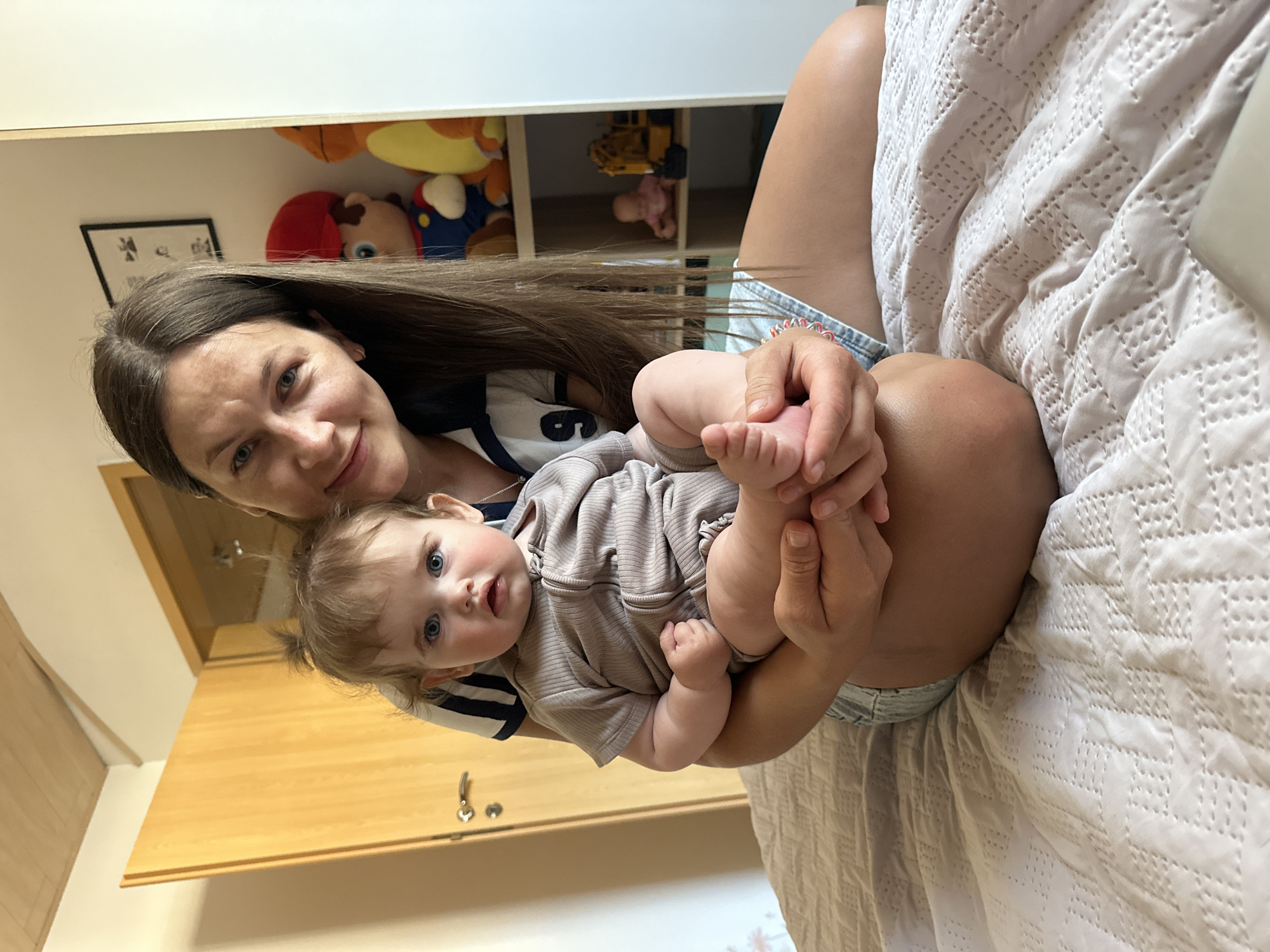 A woman is holding a baby. The woman has long brown hair and is wearing a striped shirt and jean shorts. The baby is wearing a taupe-colored romper. They are in a room with a wooden door and a shelf with toys. The scene is most likely taking place in Niederquembach, Germany, as the user requested it.