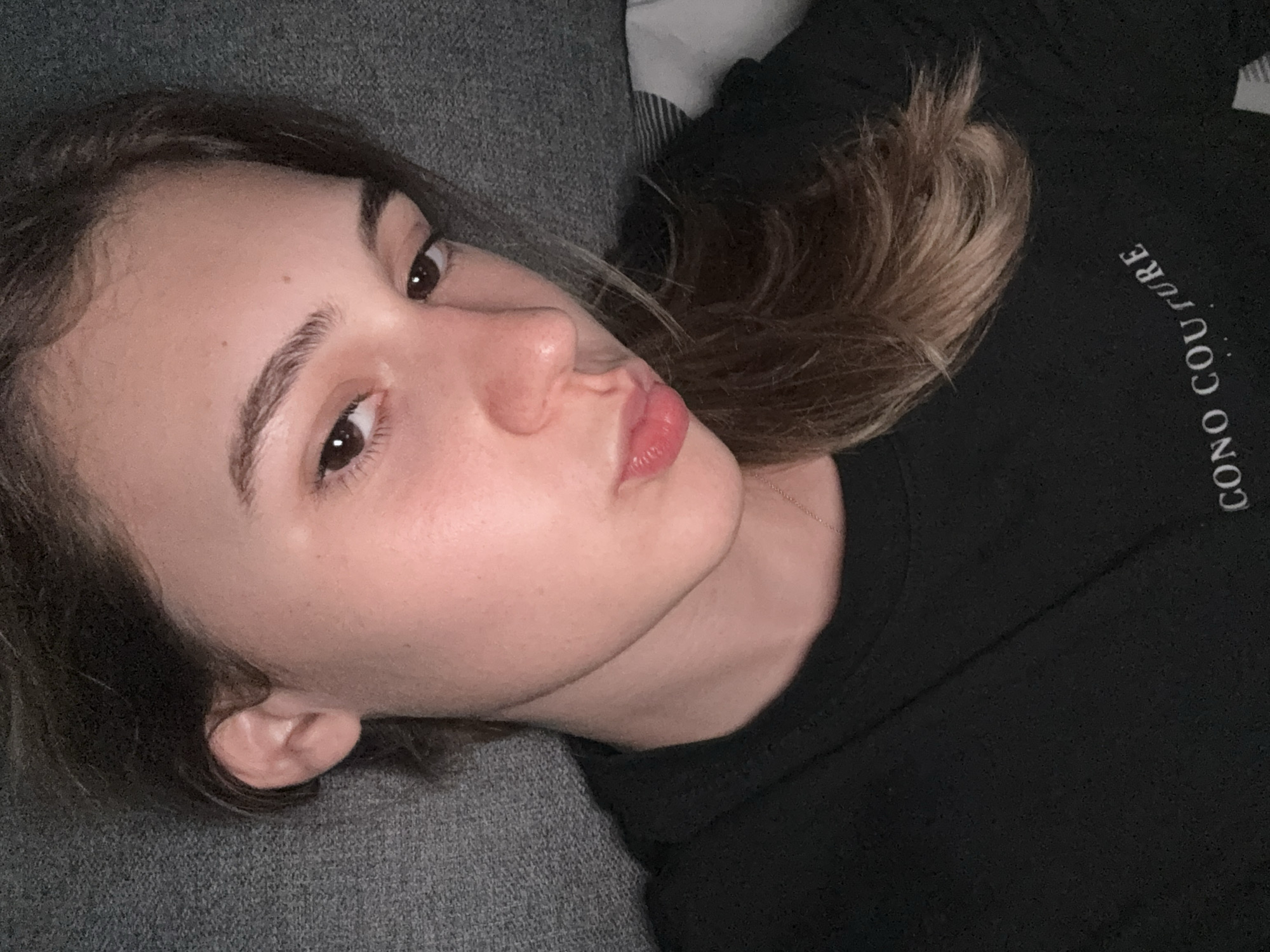 This is a selfie of a young woman lying down. She is wearing a black t-shirt with the text "CONO COUTURE" printed on it. Her head is tilted and she is looking towards the camera with her lips pursed. The background is a textured gray surface, possibly a sofa or a bed. The lighting is dim, creating a soft glow on her face. There are no identifying landmarks or clues to pinpoint the location to Forbach, Germany.