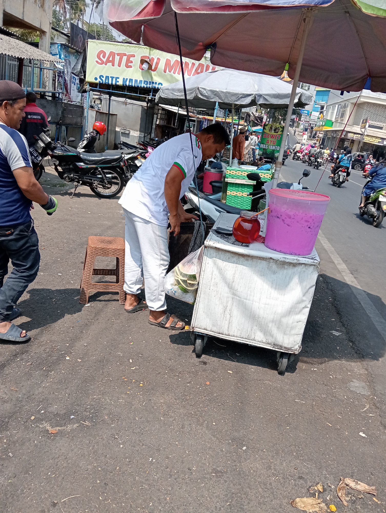 A street food vendor is operating a cart on a sunny day in Grabag, Indonesia. The cart, a white box on wheels, displays a large pink container of a beverage, a smaller glass jar of reddish liquid, and a bag of green coconuts. The vendor, wearing a white shirt and light-colored pants, is interacting with the contents of the cart. Another man, dressed in a blue shirt and dark pants, is standing to the left, looking away from the cart and the vendor. Several motorcycles are parked behind the cart, and traffic is visible in the background. A banner with Indonesian text, including "SATE GU MINA" and "SATE KAMBING," hangs above the scene, indicating the presence of satay stalls in the vicinity. The ground is paved with asphalt and shows some debris.