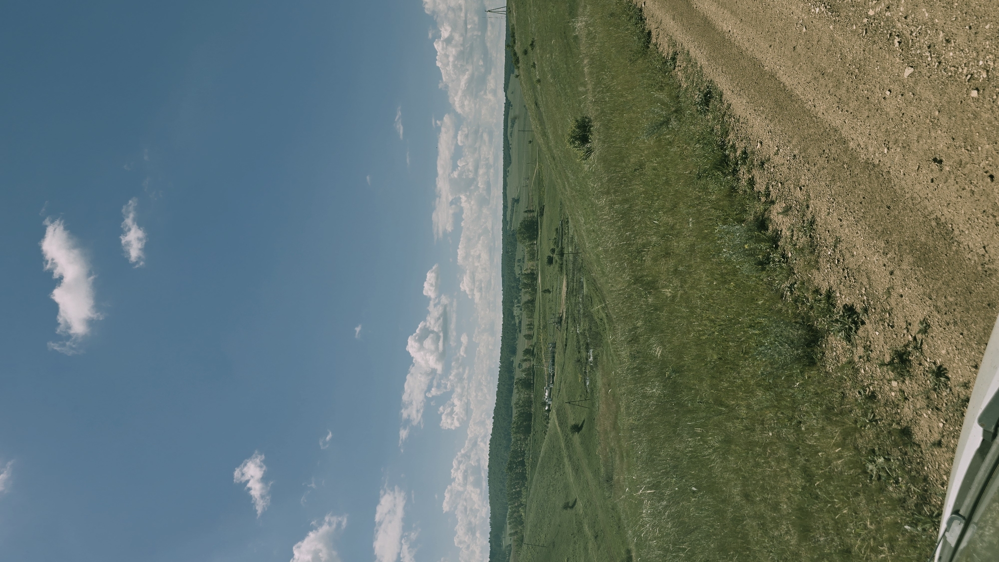 A wide-angle, eye-level shot captures a rural landscape under a clear blue sky dotted with scattered white clouds. The foreground is dominated by a dirt road curving to the right, bordered by tall, dry grass. Beyond the road, rolling green hills stretch into the distance, dotted with trees and small buildings, suggesting a village or settlement. In the far background, a dense forest forms a dark green line against the horizon. The scene conveys a sense of open space and natural beauty, typical of a countryside setting in Kudashevo, Russia. No people are visible in the image.