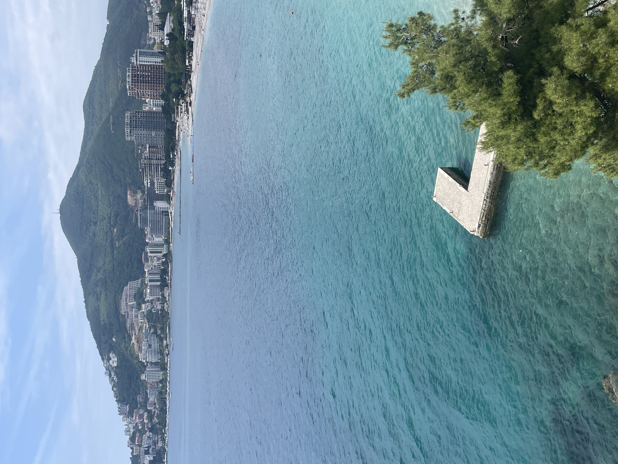 The image shows a coastline of Budva, Montenegro. High-rise buildings and houses are situated along the base of a mountain with dense green vegetation. The Adriatic Sea transitions from dark blue to turquoise near the shore. A small concrete pier extends into the sea near some trees in the right side of the frame. The sky is mostly clear.