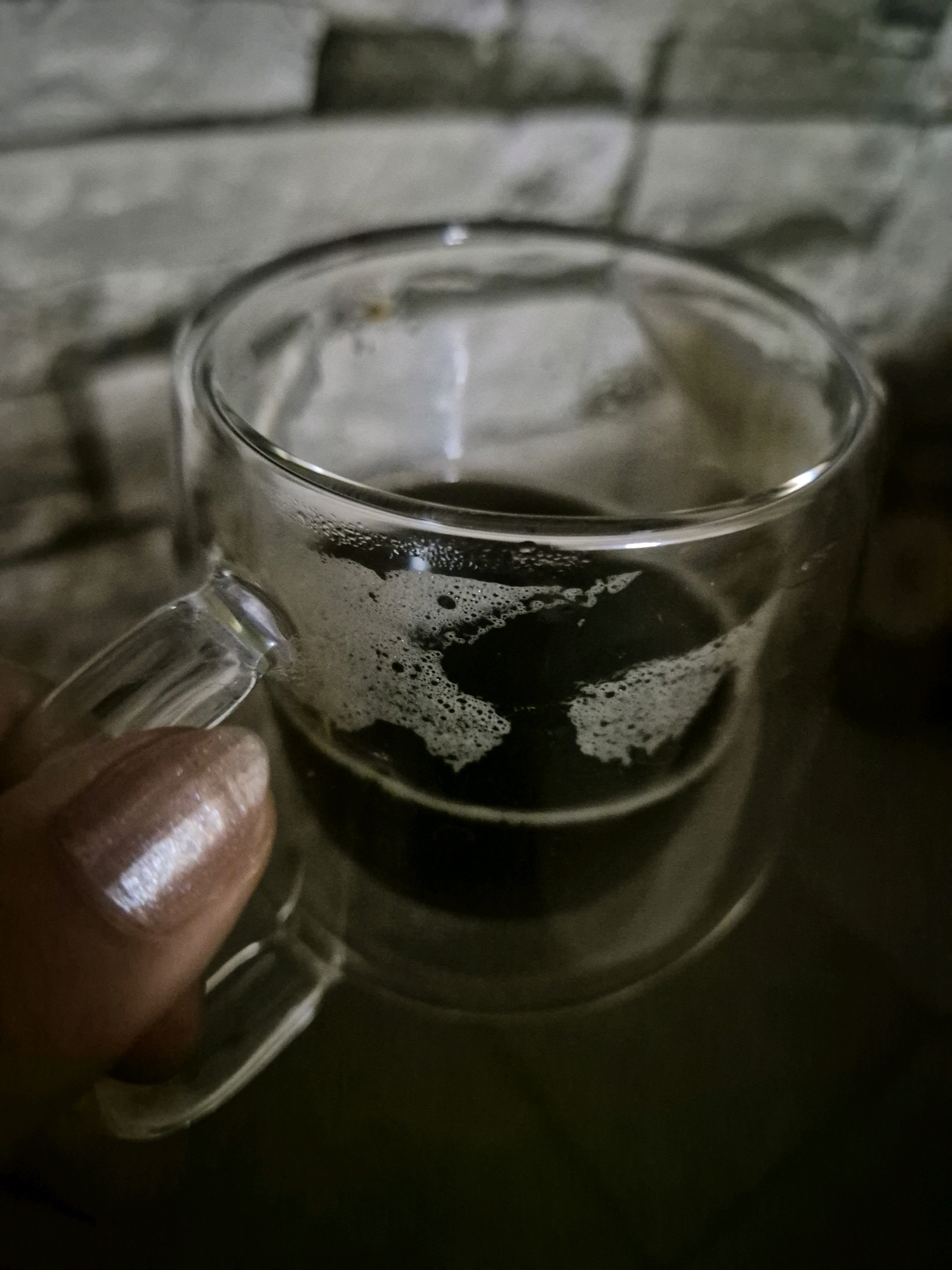 A close-up, low-angle shot shows a person's thumb and forefinger holding the handle of a clear glass mug filled with dark liquid. The mug has a double-walled design, and the surface of the liquid has a layer of foam. The background is a textured gray wall. The lighting is dim, with a strong highlight on the fingernail of the thumb.
