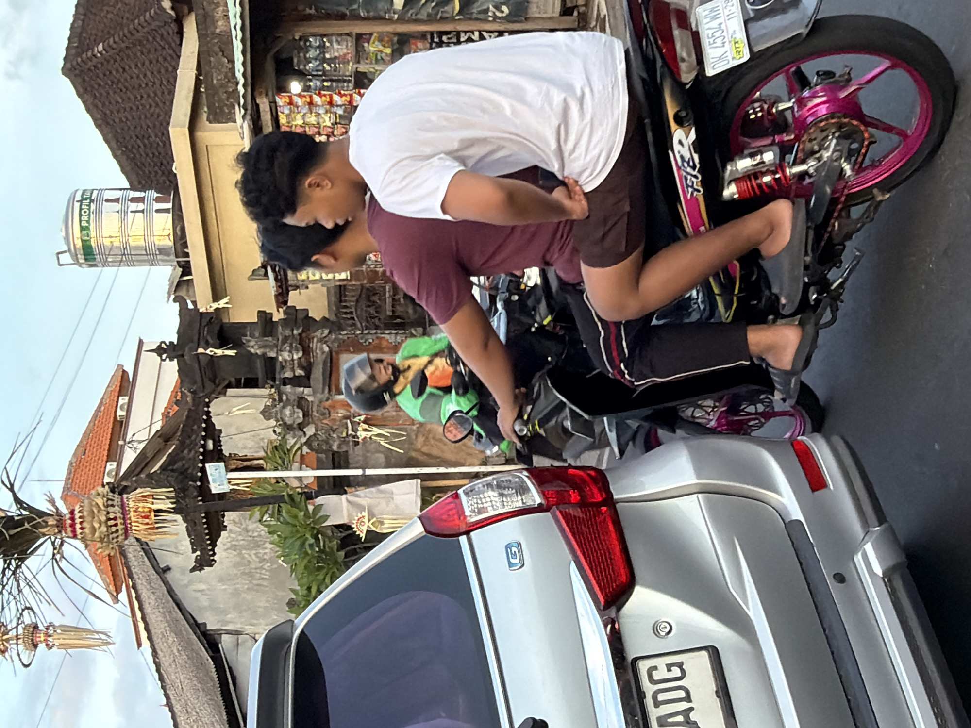 In North Kuta, Indonesia, two young men are riding a motorcycle. The man in the rear is wearing a white t-shirt and brown shorts, while the man in front is wearing a maroon t-shirt and black pants. The motorcycle has a pink wheel rim. They are positioned to the right of a silver car. In the background, traditional Balinese architecture with ornate rooflines and decorative elements is visible. A large metal water tank is mounted on a rooftop, and some overhead power lines are present. The scene appears to be on a street, possibly during the day given the lighting.