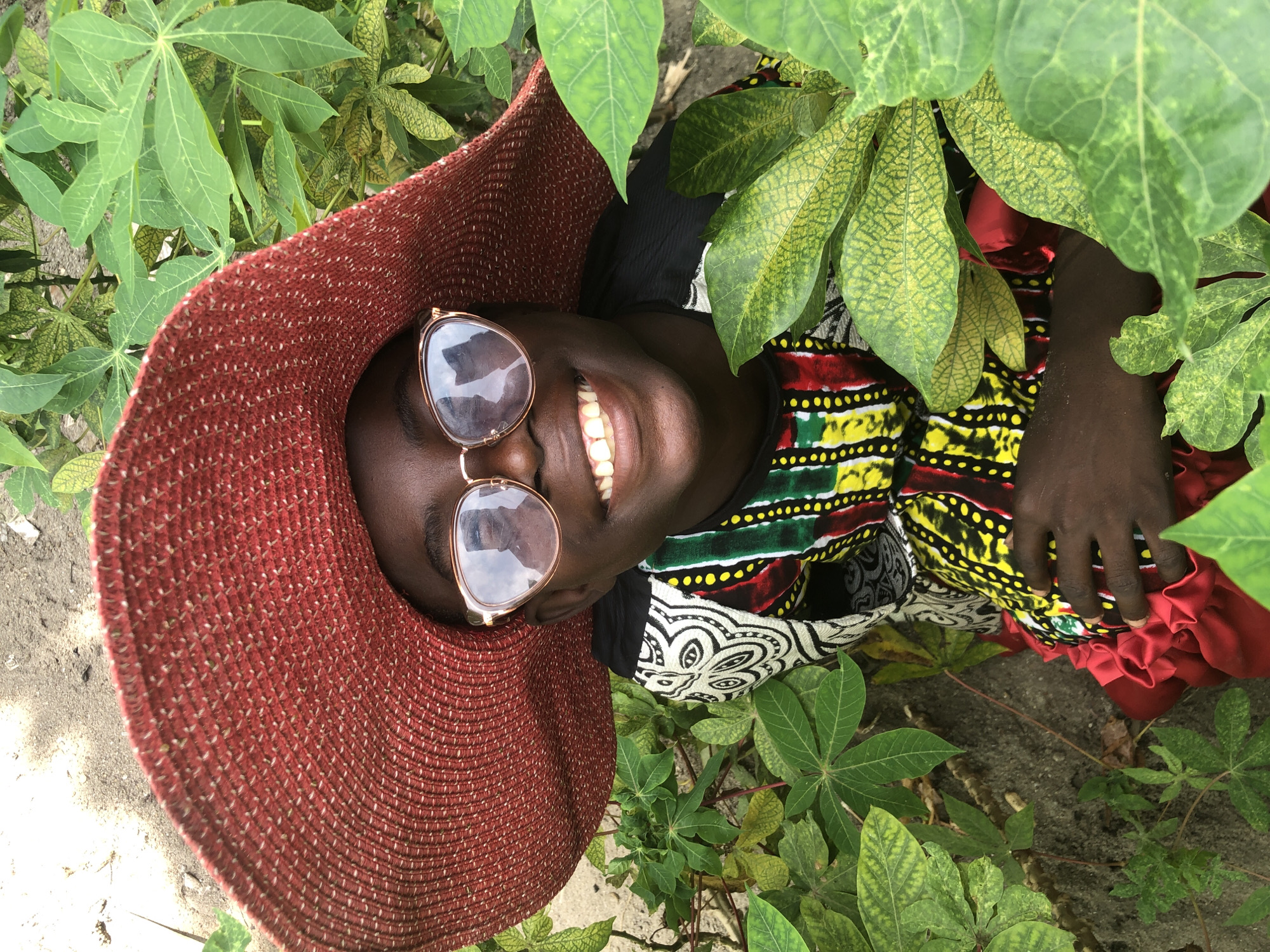 A high-angle, eye-level medium shot shows a young Black person wearing a large, red, wide-brimmed hat and round, gold-rimmed glasses. The person has dark skin, dark eyes, and bright white teeth, and is smiling. The glasses reflect a sky and possibly a ceiling with lights. The person is wearing a colorful, patterned garment that appears to be made of fabric with vibrant yellow, red, green, and black designs. The person is surrounded by lush green foliage, with some leaves showing signs of yellowing. The ground appears to be sandy or dirt. This scene is likely located in Ado, Nigeria.