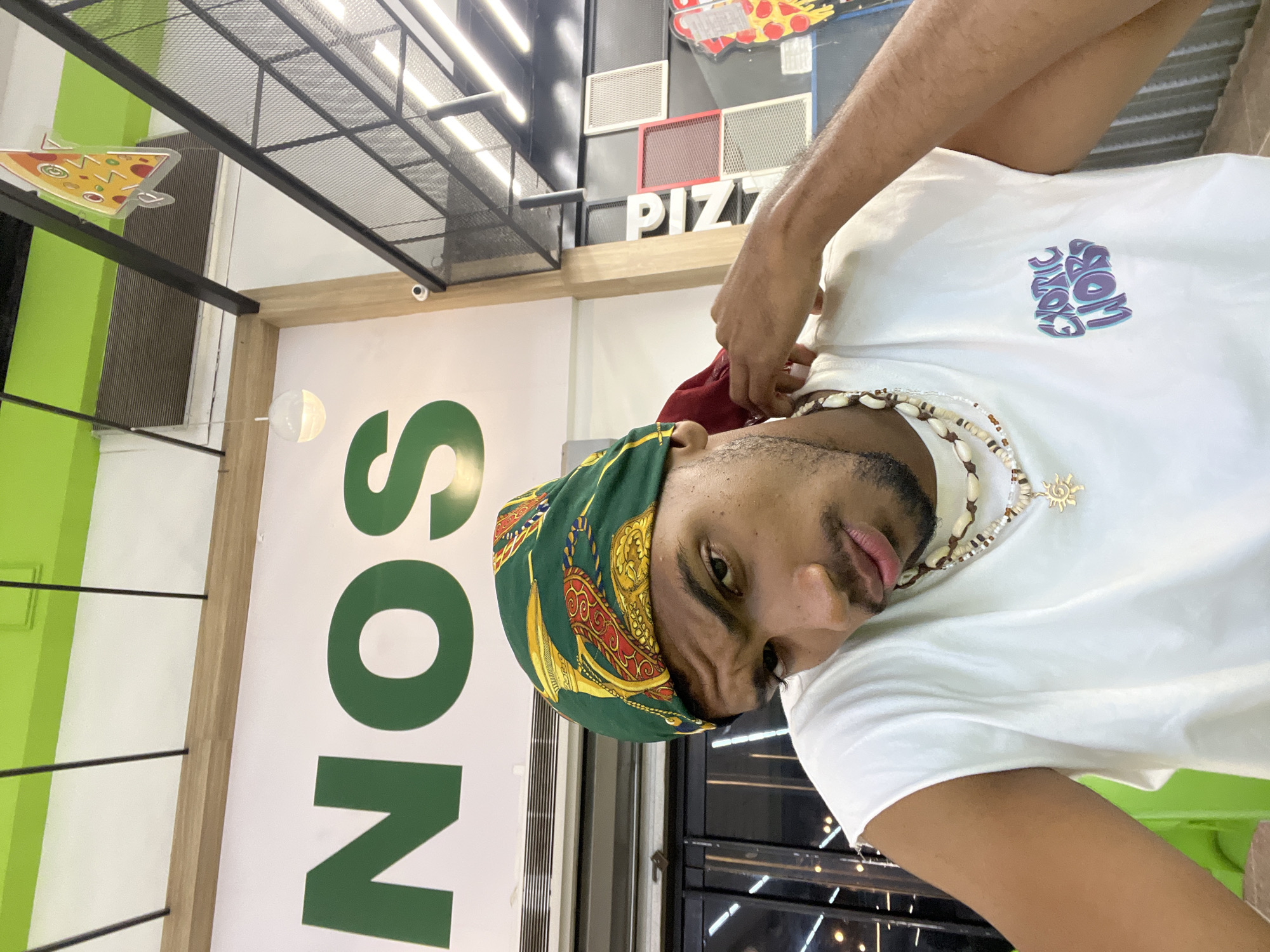 A person is taking a selfie inside what appears to be a fast-food restaurant. The sign "SON" is partially visible in the background, along with a decorative pizza slice and the word "PIZZA." The person is wearing a green and gold headscarf, a white t-shirt with a logo, and a seashell necklace. The location is likely in the city of Porlamar, Venezuela.
