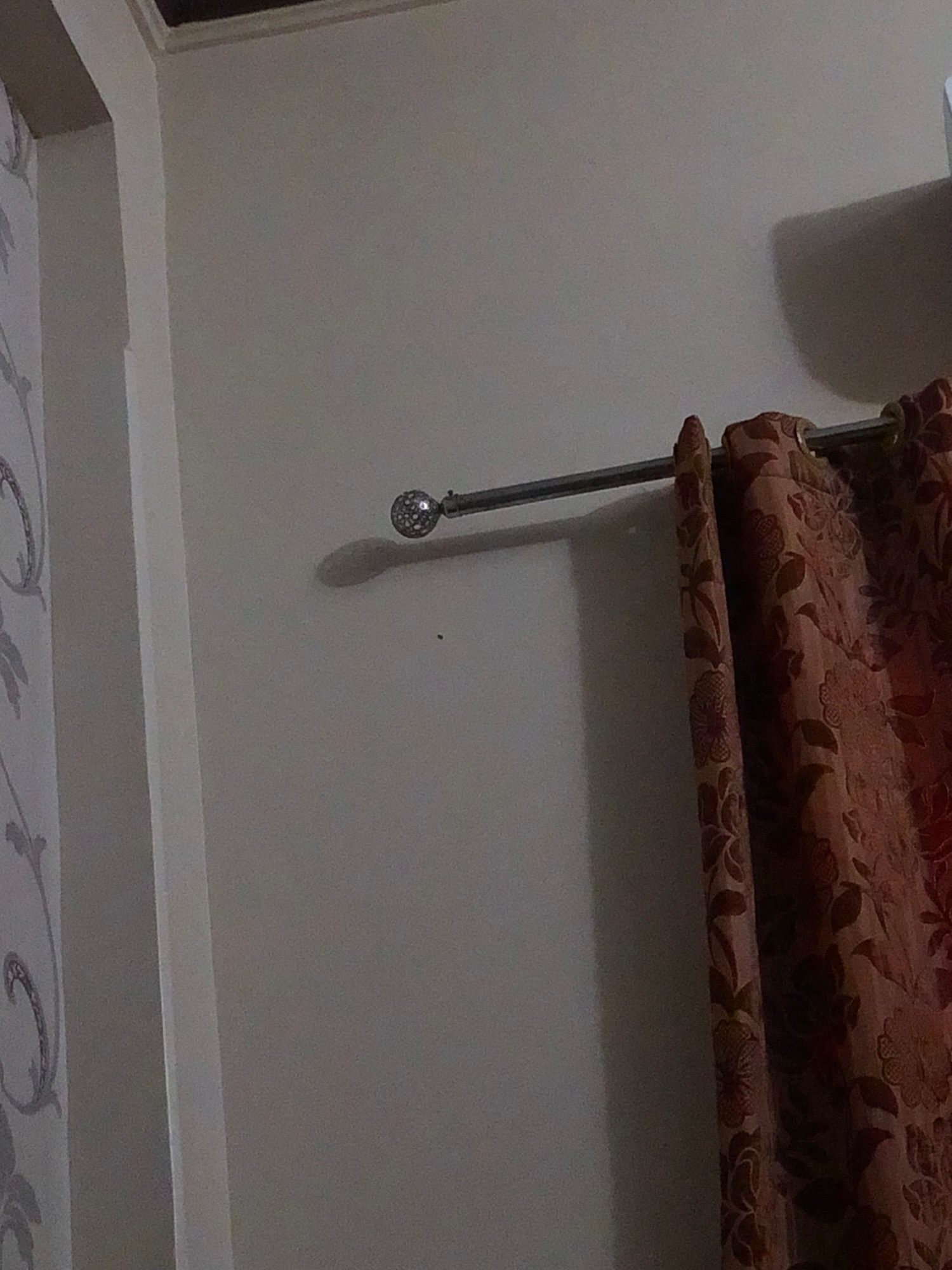 The image shows a close-up view of a wall with a curtain rod and partially drawn curtains. The wall appears to be painted a light, neutral color. A metal curtain rod extends horizontally across the image, with a decorative spherical finial at its left end. The finial is clear and appears to be faceted, reflecting light. A portion of a curtain, made of fabric with a floral pattern in shades of brown and red, hangs from the rod on the right side of the image. The curtain is drawn partially closed, obscuring the wall behind it. A shadow is cast on the wall by the curtain rod and finial, indicating a light source from the right. On the left side of the image, a portion of a wall covered in patterned wallpaper is visible. The wallpaper has a light background with a subtle, decorative design. The overall scene suggests an indoor setting, likely a room in a residential or commercial building. No people or specific actions are depicted. There is no indication of the location being in Larkana, Pakistan.