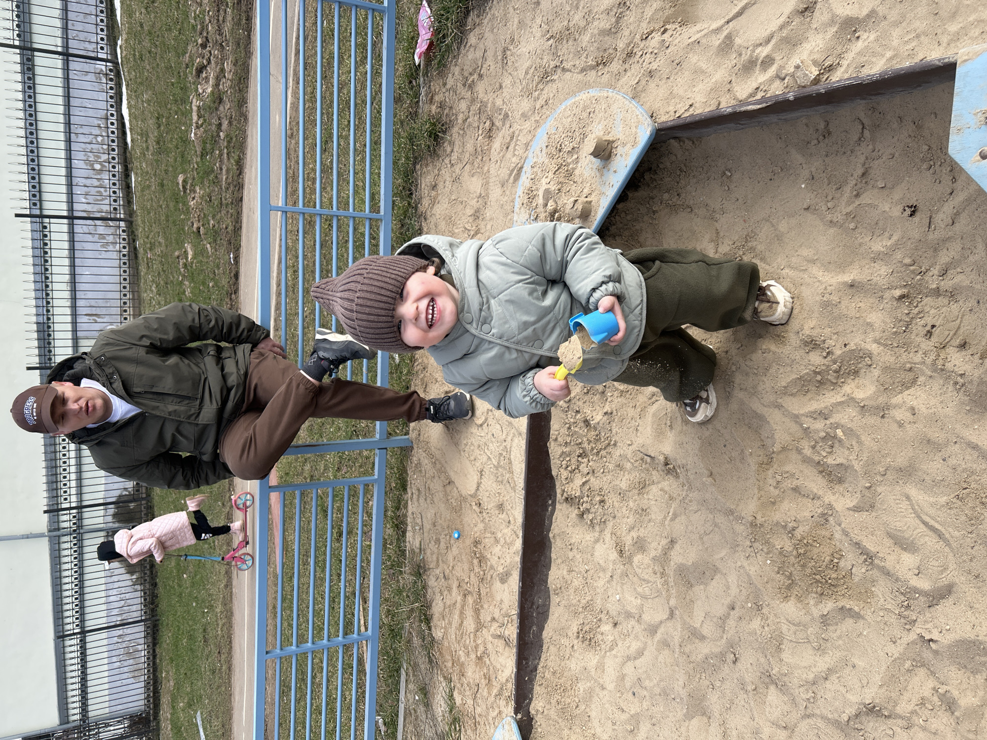 Observed in Klin, Russia, the image captures a playground scene. A young child, donning a brown knit hat and a light green quilted jacket, stands in a sandbox. The child is holding a blue cup filled with sand and a yellow toy shovel, displaying a broad smile with visible teeth. Adjacent to the sandbox, an adult male is seated on a blue metal fence, wearing a brown baseball cap, a dark green jacket, and brown trousers. Further in the background, partially obscured by the fence, a smaller child is riding a pink scooter. The sandbox itself is constructed with wooden and blue metal elements. The environment surrounding the playground includes a grassy area and a building in the distance.