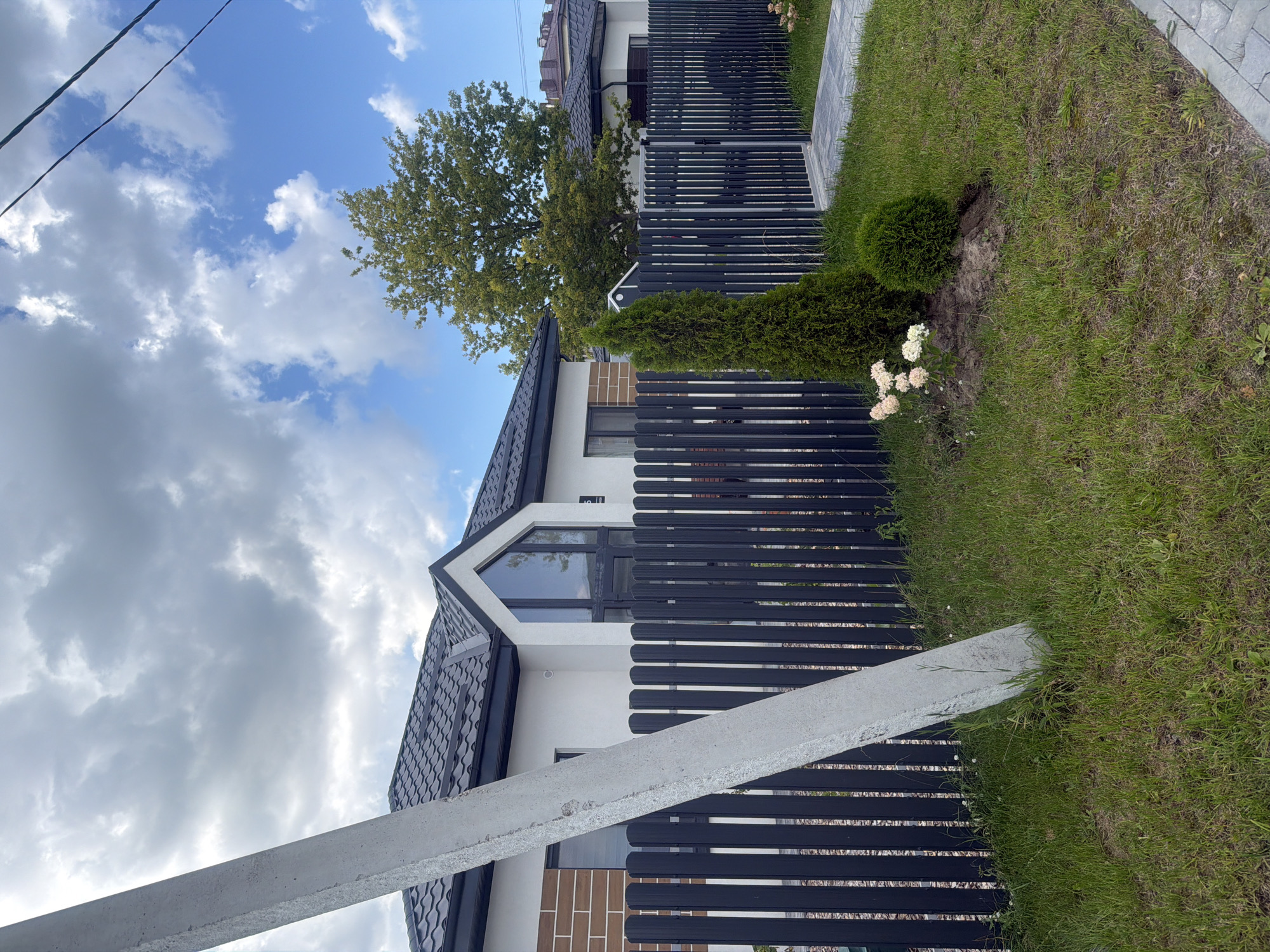 The image depicts a modern, low-rise house with a dark-tiled roof and white walls, partially accented with brickwork. The property is enclosed by a dark, vertical-slatted fence with a gate. In the foreground, a large, gray concrete utility pole is positioned at an angle, leaning towards the left side of the frame. The yard is covered in green grass, with a small evergreen tree and a rounded shrub planted near the fence. Several light-pink flowers are also visible in the garden bed. The sky is partly cloudy with patches of blue. The house is situated in the city of Golubevo, Russia.