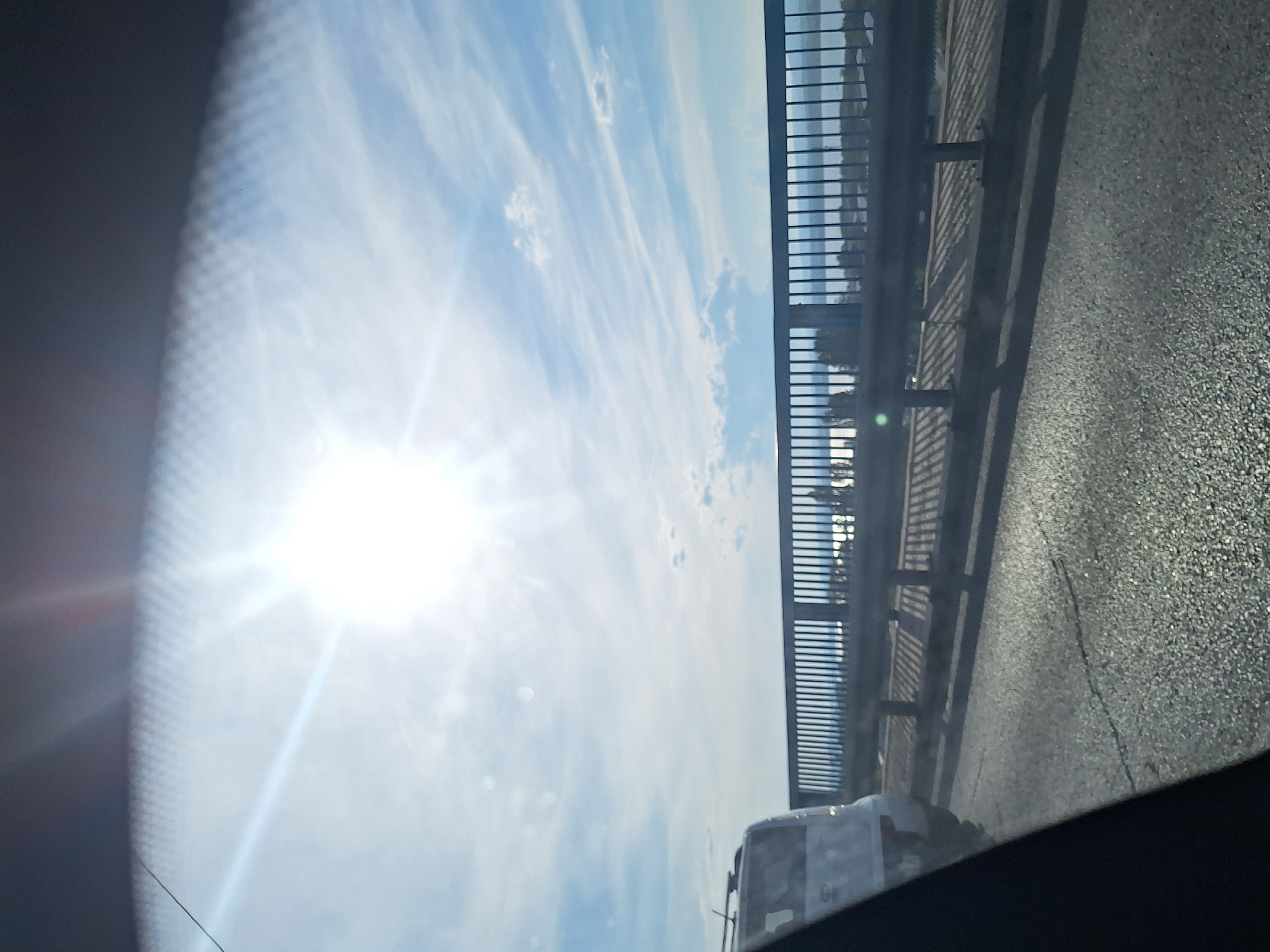 The image captures a bright, sunny day viewed from inside a vehicle, likely in Varna, Bulgaria. The sun is high in the sky, creating lens flare and washing out much of the detail in the upper portion of the image. A light blue sky with scattered white clouds stretches across the horizon. In the middle ground, a metal guardrail with vertical bars runs horizontally across the frame, casting long shadows onto the asphalt road below. Beyond the guardrail, a body of water is visible, reflecting the sunlight, with distant landmass and trees on the other side. To the left of the guardrail, the rear of a white SUV is partially visible, indicating the vantage point is from another moving vehicle. The road surface is asphalt with visible cracks and a gritty texture. The overall impression is of a scenic drive on a clear, sunny day, likely near a coastal area given the presence of a large body of water.