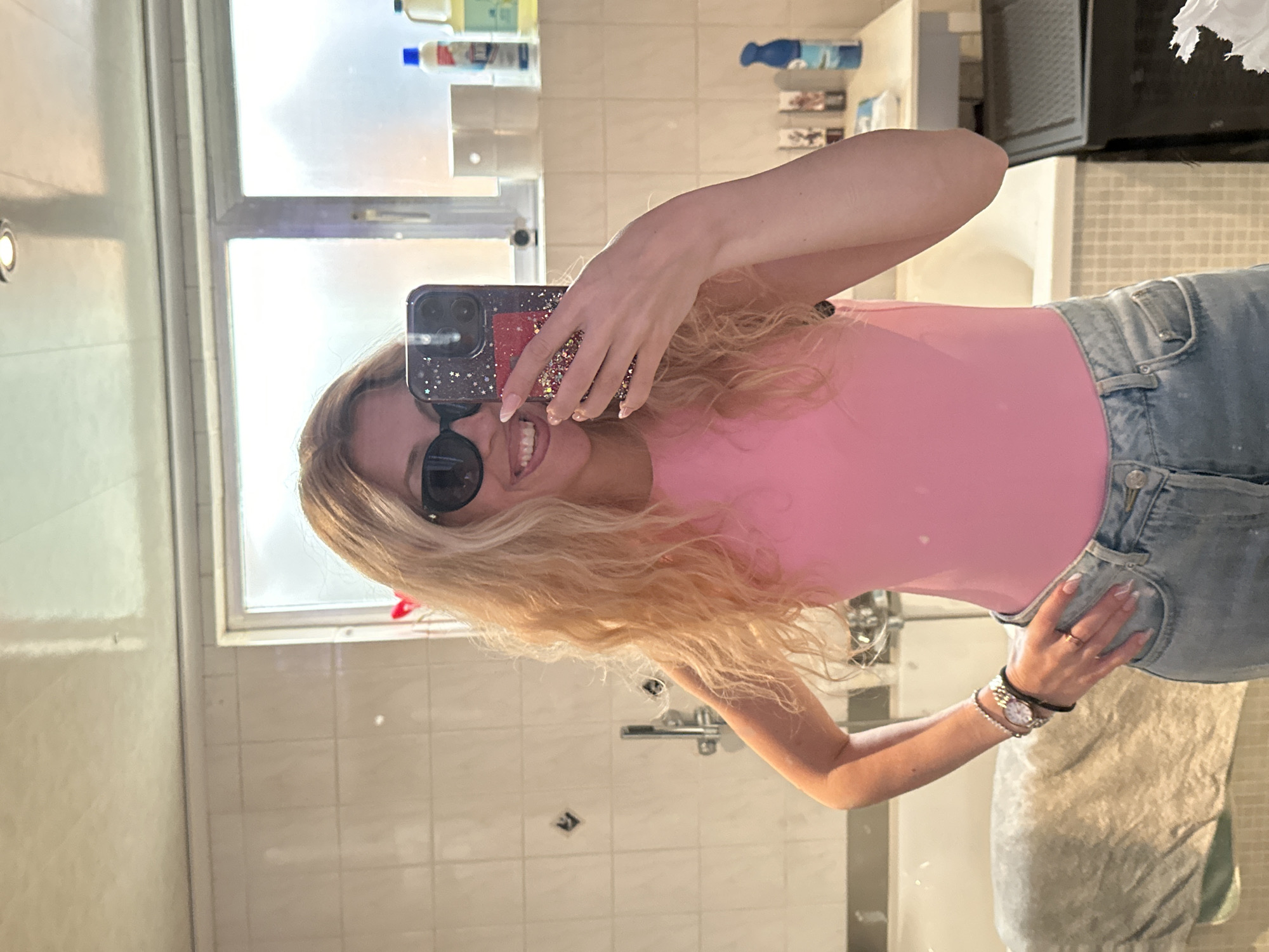 A young woman with blonde, curly hair, wearing black sunglasses, a pink sleeveless top, and light-wash denim jeans, is taking a mirror selfie in a bathroom. She is smiling and holding a phone decorated with stars and glitter. She is also wearing a watch on her left wrist and rings on her fingers. The bathroom has tiled walls, a bathtub, and shelves with various bottles and containers. The overall scene appears to be a private residence. There is no indication that this image was taken in Bruges, Belgium.