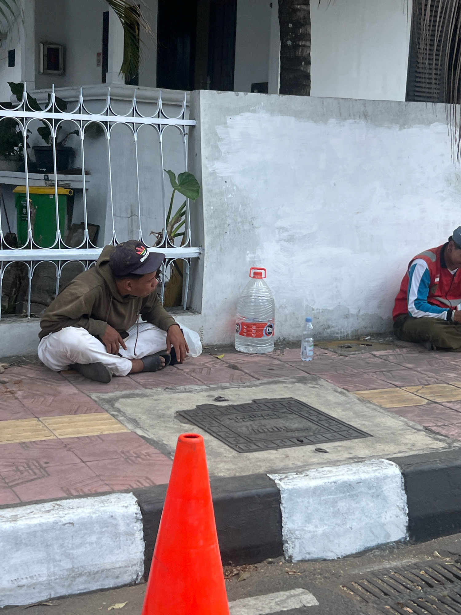 A young man is sitting on the sidewalk in front of a white building, resting his head on his hand and looking to the right. He is wearing a brown hooded sweatshirt, white pants, and dark sandals. A large bottle of water and a smaller bottle of water are on the ground next to him. In the background, another person is sitting on the ground, leaning against the wall, wearing a red vest. A white fence is visible to the left of the young man. A red traffic cone is in the foreground on the right. The scene appears to be on a sidewalk in front of a building, possibly in a public area within Yogyakarta, Indonesia.