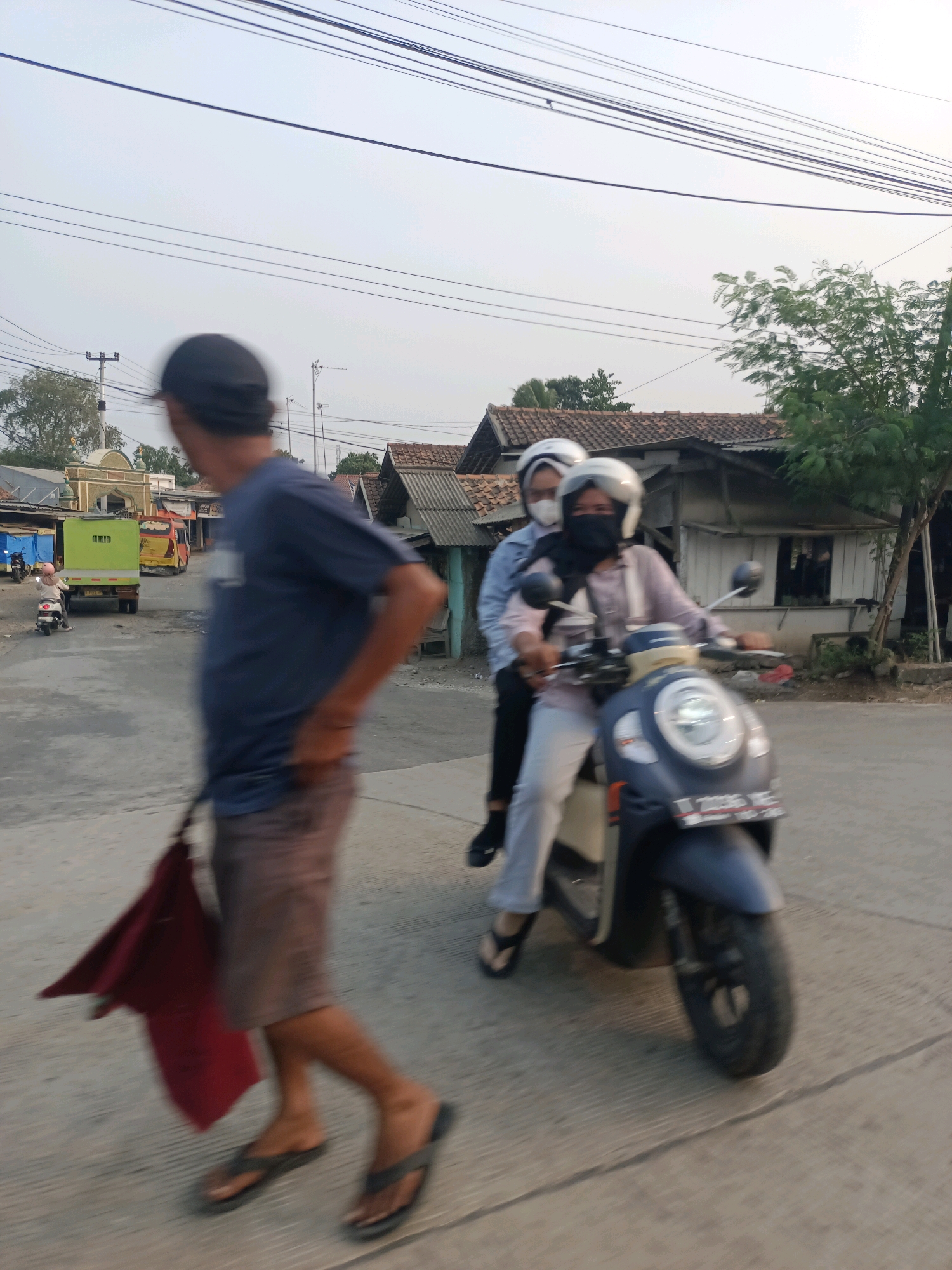 The image depicts a street scene in Subang, Indonesia, with a focus on motion blur, suggesting the photo was taken while moving or of moving subjects. In the foreground, a person wearing a dark cap and a navy blue t-shirt is walking towards the right, carrying a red cloth bag in their left hand. They are wearing brown shorts and dark flip-flops. To the right of the foreground figure, a dark blue and cream-colored scooter is moving away from the camera, carrying two individuals. Both passengers are wearing helmets and face coverings. The driver, in the front, is wearing a light purple long-sleeved shirt and white pants. The passenger behind them is wearing a light blue long-sleeved shirt. The background shows residential buildings with tiled roofs and some greenery. Power lines are visible overhead. A lime green truck and a yellow truck are parked or moving further down the street.