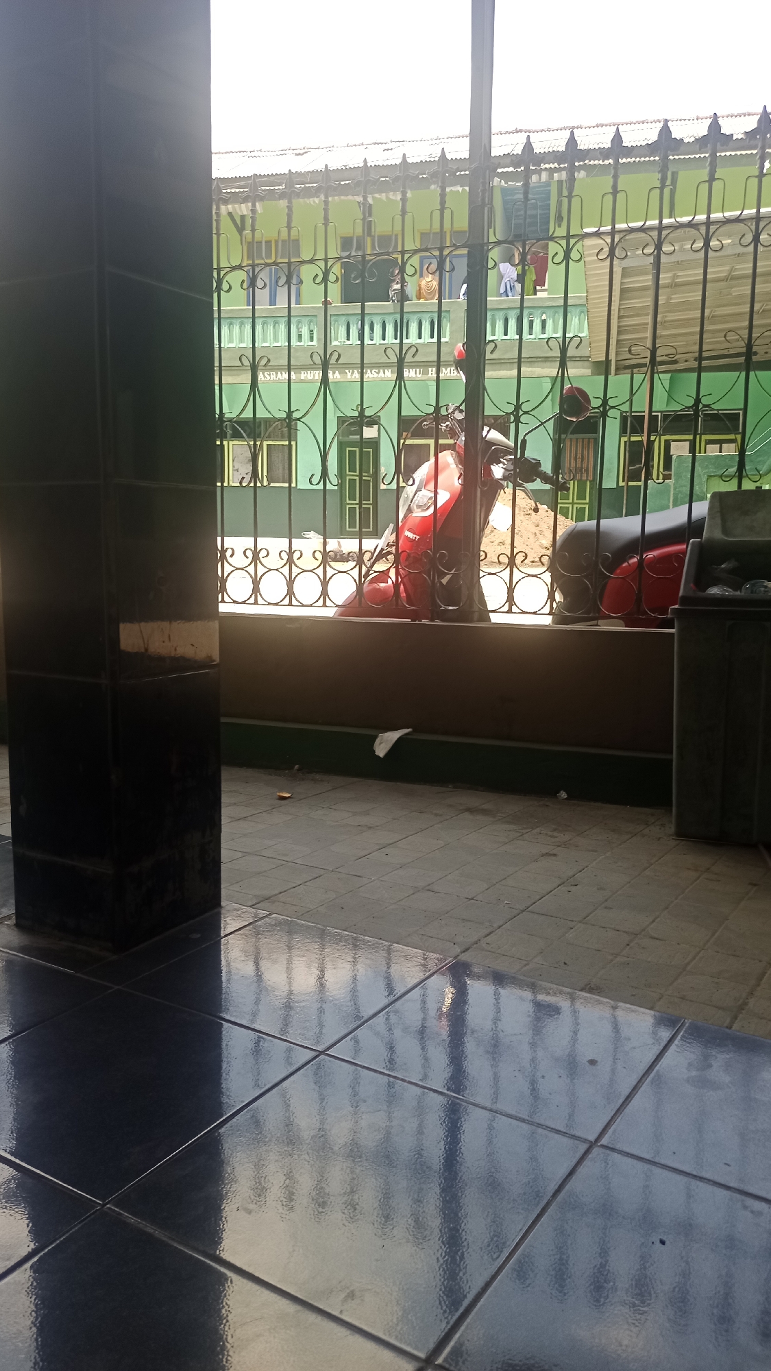 The image depicts a scene from Karyamandala, Indonesia, viewed through a metal fence. In the foreground, a dark, tiled floor reflects the environment. A large, dark, rectangular pillar is present on the left side of the frame. Beyond the fence, a red scooter is parked. Behind the scooter, a green building with multiple windows and a covered walkway is visible. The building has signage that reads "ASRAMA PUTRA YA'ASAN BNU H ME". Laundry is hanging on a line on the upper level of the building. The overall impression is of a street-side view of a building complex.