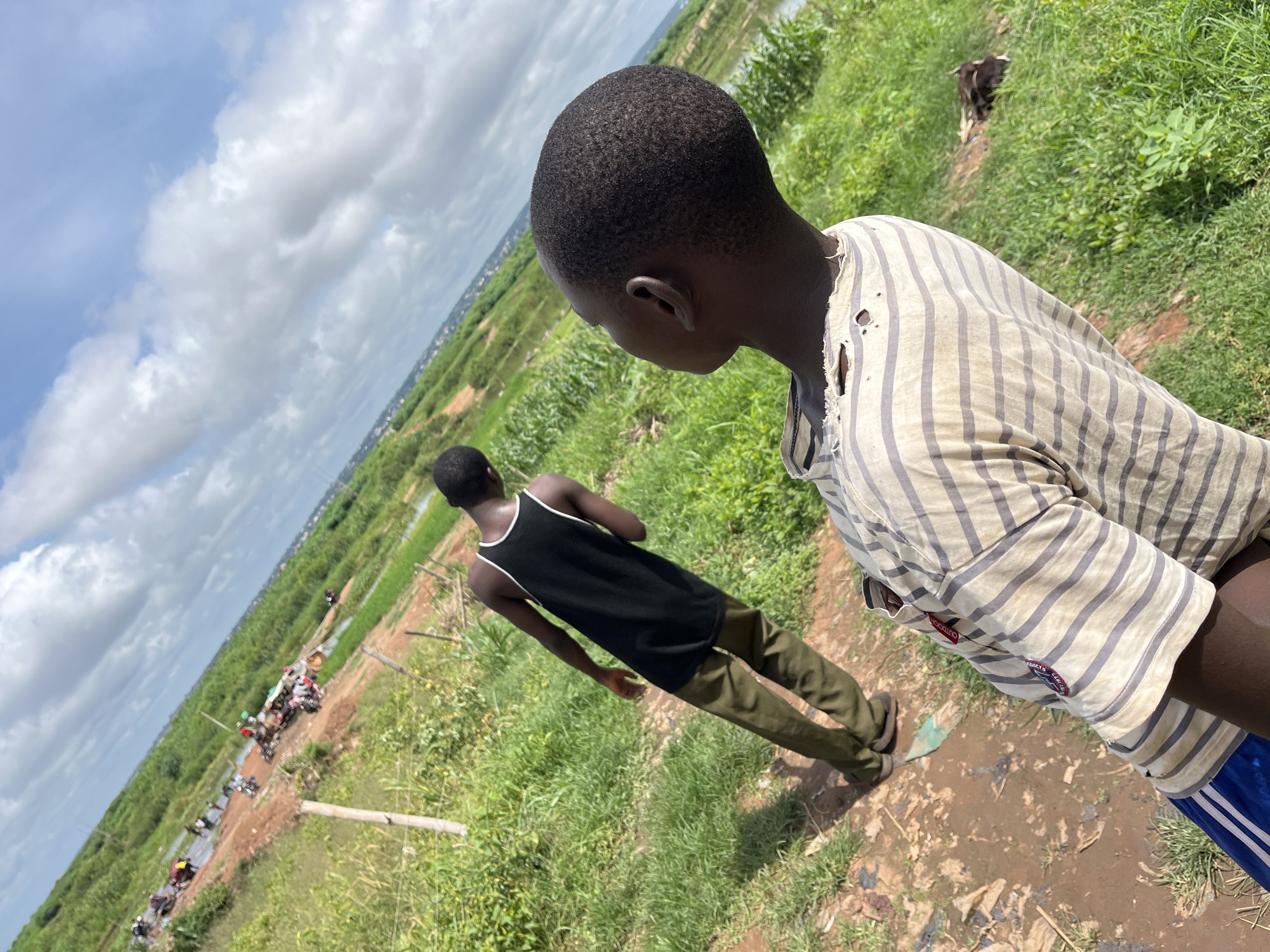 This image presents an outdoor scene in Idah, Nigeria, featuring two young males amidst a vibrant green landscape under a bright, partly cloudy sky. The primary subject is a young male seen from the back and side in the foreground, with short dark hair and dark skin. He is wearing a faded, off-white long-sleeved shirt with grey stripes, which shows signs of wear with several visible holes. His lower attire consists of blue shorts with distinct white stripes. On the sleeve of his shirt, a small red tag with white text reading "OUTDOOR" is visible, along with another smaller blue tag with partially obscured white text, likely "PRODUC" or "PRODUCTS".
Further into the mid-ground, a second young male with dark skin is seen from behind, walking away from the camera. He is dressed in a black tank top with white trim around the neck and armholes, paired with olive green pants. Both individuals appear to be walking along a rough, muddy dirt path that cuts through extensive, lush green vegetation, possibly crops or tall grasses.
In the background, a long, winding dirt track or road can be seen, lined with several motorbikes or motorcycles, suggesting activity or a settlement in the distance. A body of water, perhaps a river or a large ditch, runs parallel to the green expanse. In the upper right background, a dark animal, possibly a goat, is partially visible among the greenery. The sky is bright blue with large, white cumulus clouds, indicating it is daytime. The overall scene conveys a rural or semi-rural environment.