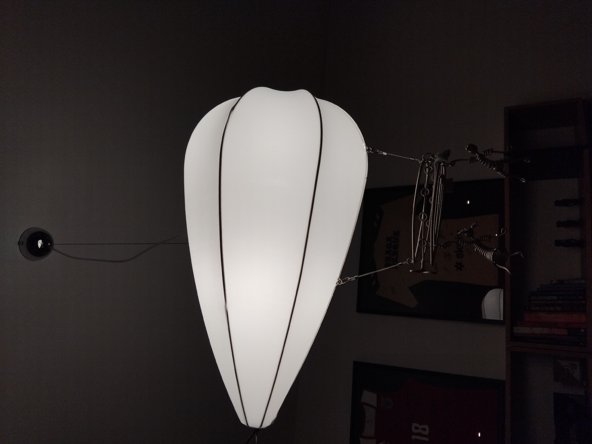 The image shows a close-up of a modern, white, balloon-shaped pendant light fixture hanging from a dark ceiling. The light fixture is suspended by a thin wire and has two black bands circling its circumference. Attached to the bottom of the light fixture is a decorative element resembling a metal rack with two small, articulated metal figures hanging from it. In the background, slightly out of focus, are two framed posters or jerseys hanging on a wall, one with the number "18" visible. Below these posters, a wooden shelf unit is visible, containing books and other decorative items. The scene appears to be indoors, possibly in a home or retail space. The lighting is dim, with the primary light source being the pendant fixture itself.
While the image is of an interior scene, there is no specific visual information within the image to determine its location in Vila Nova de Famalicão, Portugal. The description of the scene is based solely on the visual elements present in the photograph.