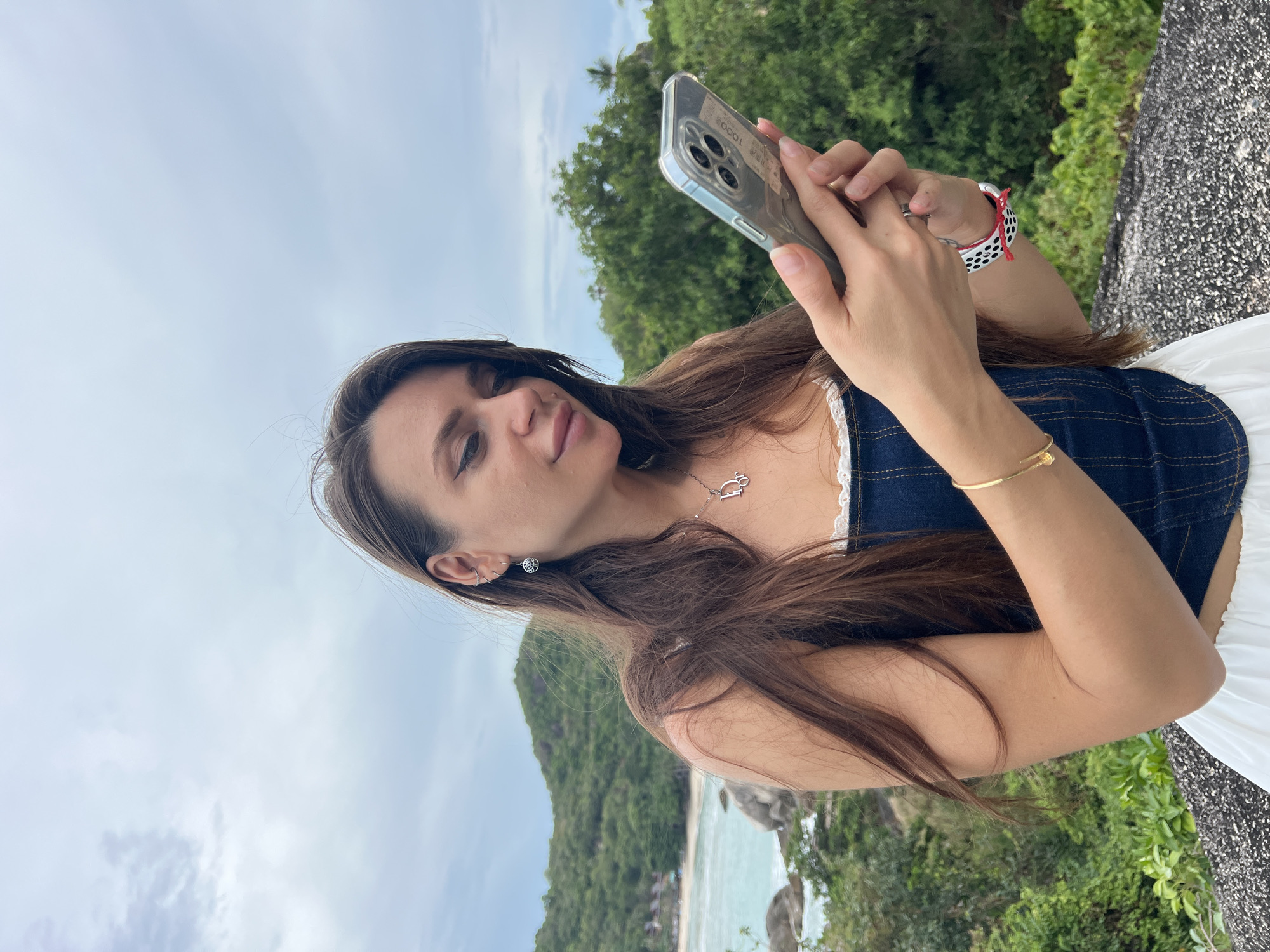 A woman stands in Baan Chaweng Noi, Thailand, holding a smartphone. The phone is clear-cased, and she is actively using it. She is wearing a denim top with white lace trim, a white skirt, and a gold bracelet. A silver necklace with a "D" pendant is visible. Her hair is long and brown. In the background, there is a view of the beach, ocean, and lush green landscape. The sky is overcast. She has an Apple Watch on her left wrist.