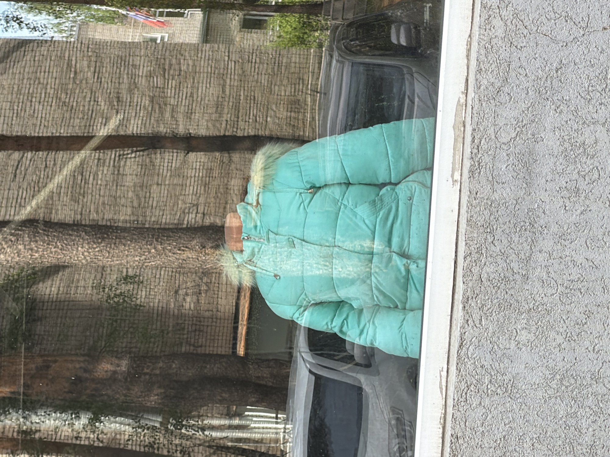 The image shows a light blue puffer jacket with fur trim hanging in a window. The reflection in the window reveals a brick building with trees. A car is also visible in the reflection. The photograph was taken during the daytime in what appears to be an urban setting. Based on the surrounding imagery, the jacket might be present in the city of Chaikovsky, Russia.