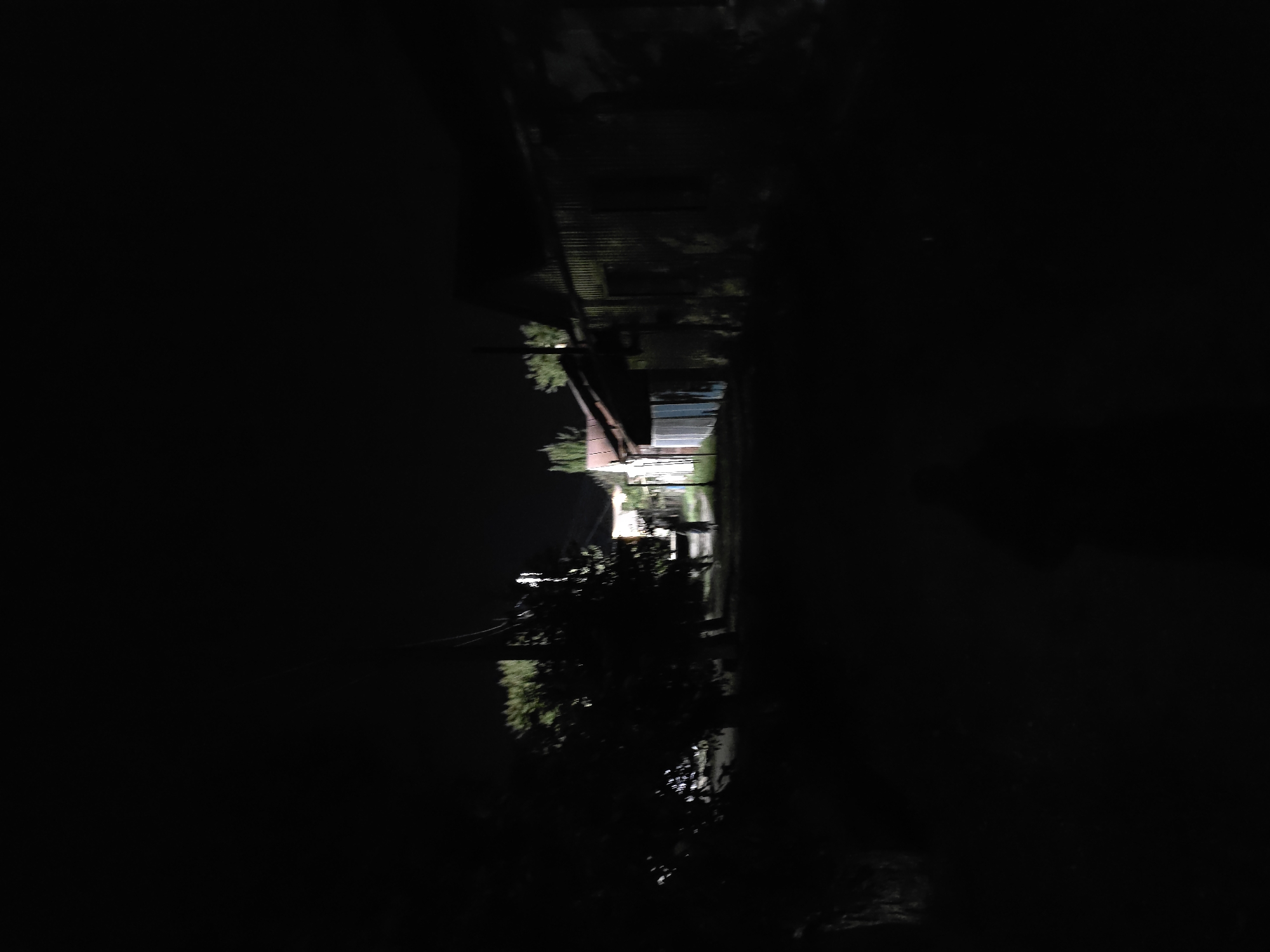 The image depicts a dimly lit outdoor scene at night, possibly in the city of Qarabalıq, Kazakhstan. On the right side of the image, a corrugated metal building with a sloped roof is visible. To the left of this building, a smaller structure with a lighter colored roof and walls is present. A person, appearing as a silhouette, is walking away from the camera in the center of the frame, on a dirt path. Trees and foliage are present on the left side of the path, casting shadows. The overall environment suggests a rural or semi-rural setting with basic structures and vegetation under the cover of darkness.