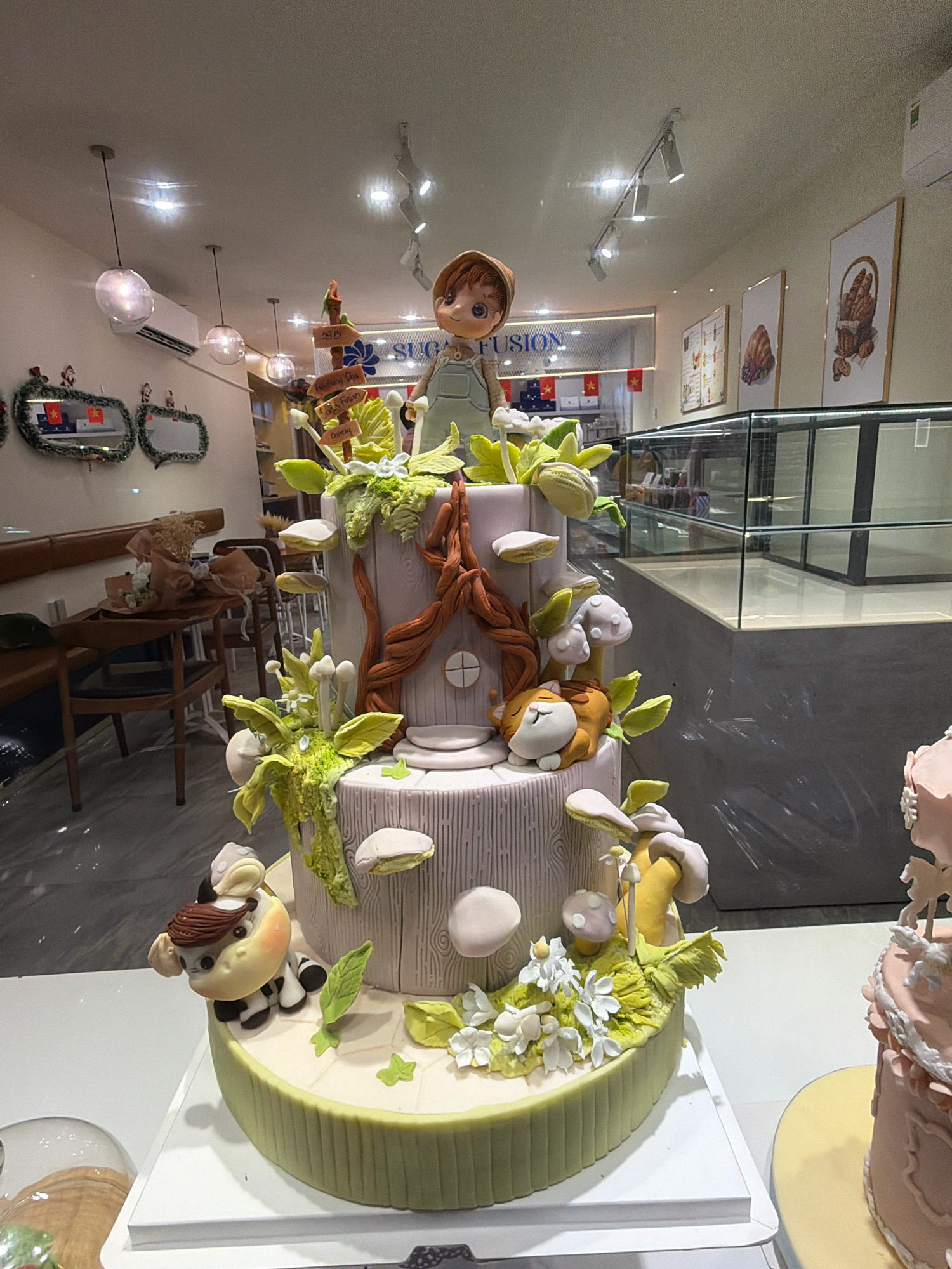 The image shows a multi-tiered cake decorated with a whimsical, fairytale theme, possibly a woodland setting. The cake is adorned with fondant figures including a child, a sleeping cat, and a cow, as well as mushroom and floral decorations. The cake is displayed in what appears to be a bakery or dessert shop. In the background, there are display cases with other desserts, framed artwork depicting baked goods, and ambient lighting from ceiling fixtures and possibly a display case. There are also mirrors with festive decorations on the wall. The overall environment suggests a commercial establishment focused on sweets. The photograph was taken in Phú Quốc, Vietnam.