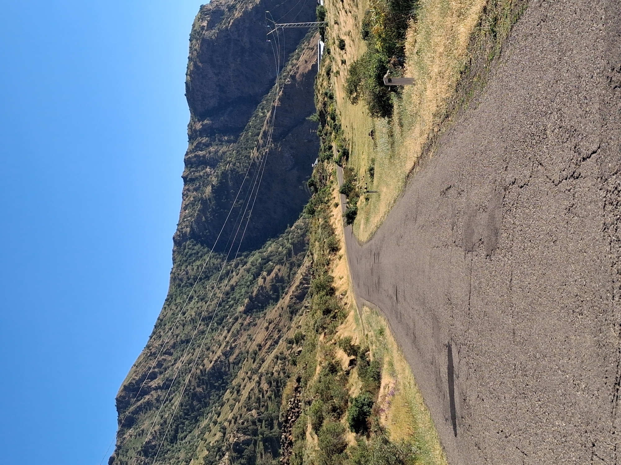 The image depicts a two-lane paved road winding through a mountainous landscape. The road surface is cracked and worn, with a dark stain visible on the left side. The terrain flanking the road is covered in dry, yellow grass and sparse green bushes. In the background, a steep, rocky mountain slope rises, dotted with green vegetation. Power lines stretch across the mountain face, supported by poles. A distant building with a white roof and a transmission tower are also visible on the right side of the frame. The sky is clear and blue. The scene appears to be located in a rural, mountainous area, likely within or near Shatin, Armenia, given the context provided. No people or animals are visible in the image.