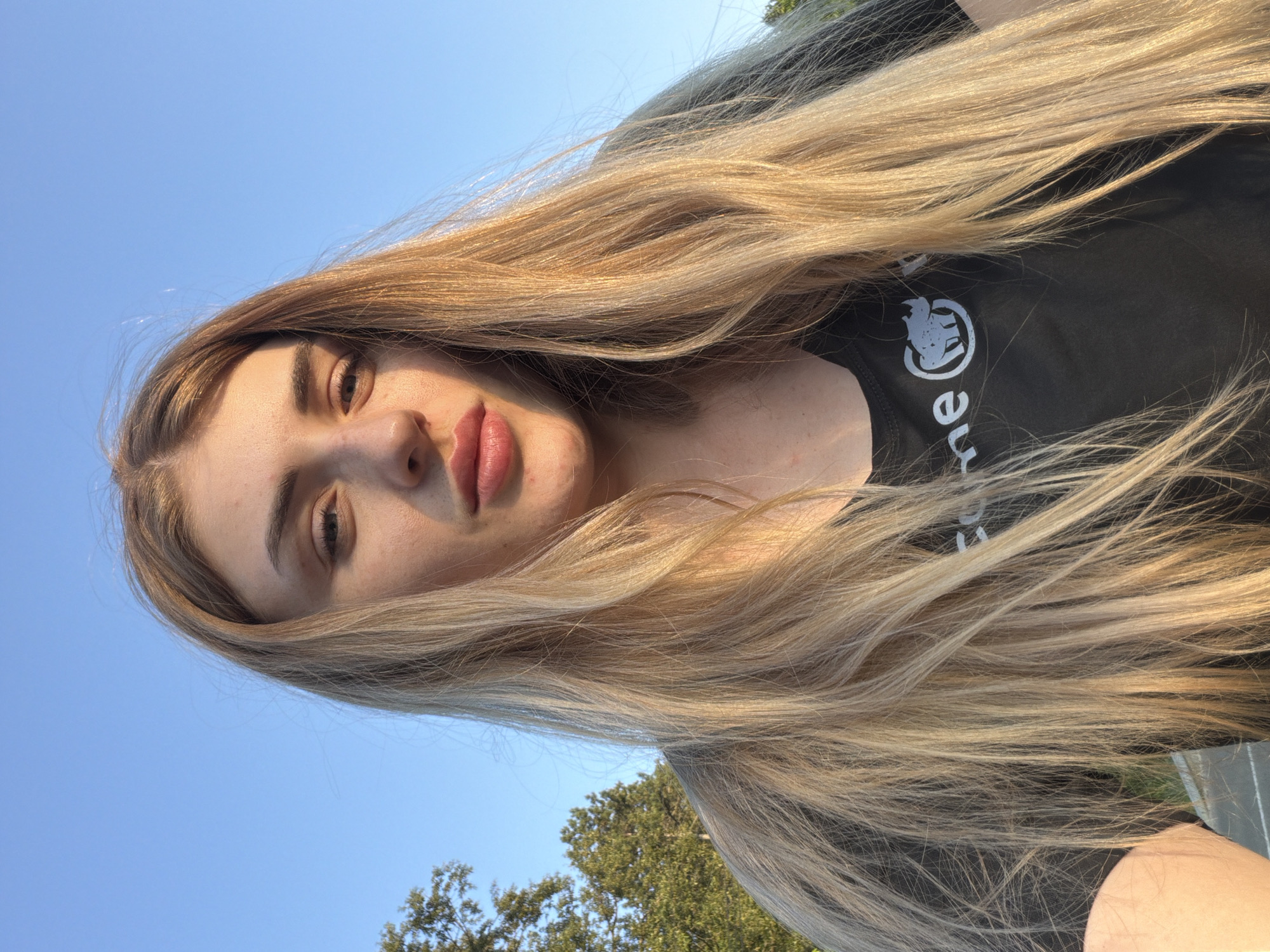 A young woman with long blonde hair is looking at the camera, outdoors against a clear blue sky. She is wearing a black t-shirt with a white graphic of a rhinoceros in a circle. The sun is casting a shadow across her face. Trees are visible in the background.