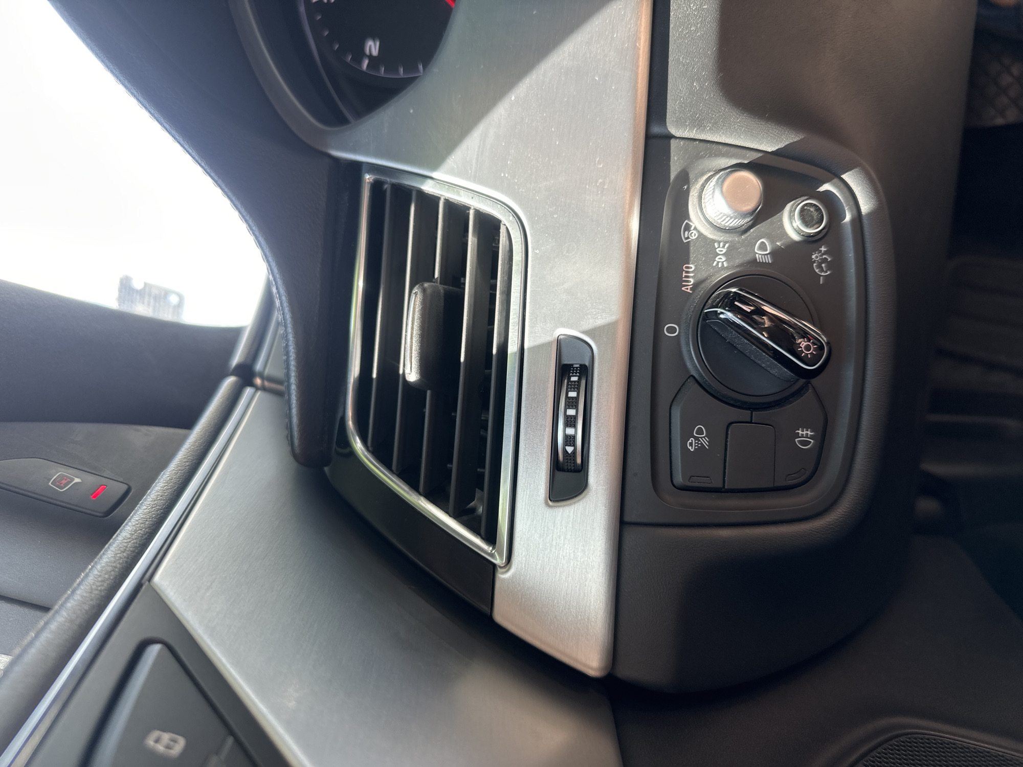 The image shows a close-up view of the interior of a car, specifically focusing on the dashboard and driver's side controls. In the foreground, a black dial with chrome accents is visible, likely for controlling headlights or interior lighting. Above this dial, a silver vent with horizontal slats is present, a common feature in car interiors. To the left of the vent, a black button with a symbol resembling a locked door is partially visible, possibly a door lock or unlock button. To the right of the dial, there's a metallic rotary knob and a smaller black button, suggesting controls for functions like fog lights or dimmer settings. A red warning light is visible on a black panel in the upper portion of the image, which is likely part of the car's dashboard or center console. The lighting in the image suggests it was taken during the daytime, with bright sunlight casting shadows on the dashboard. The environment is clearly an indoor setting, within a vehicle. There are no people or animals visible in the image. The photograph does not contain any identifiable landmarks or features that would allow for pinpointing its location within the city of Ruse, Bulgaria.