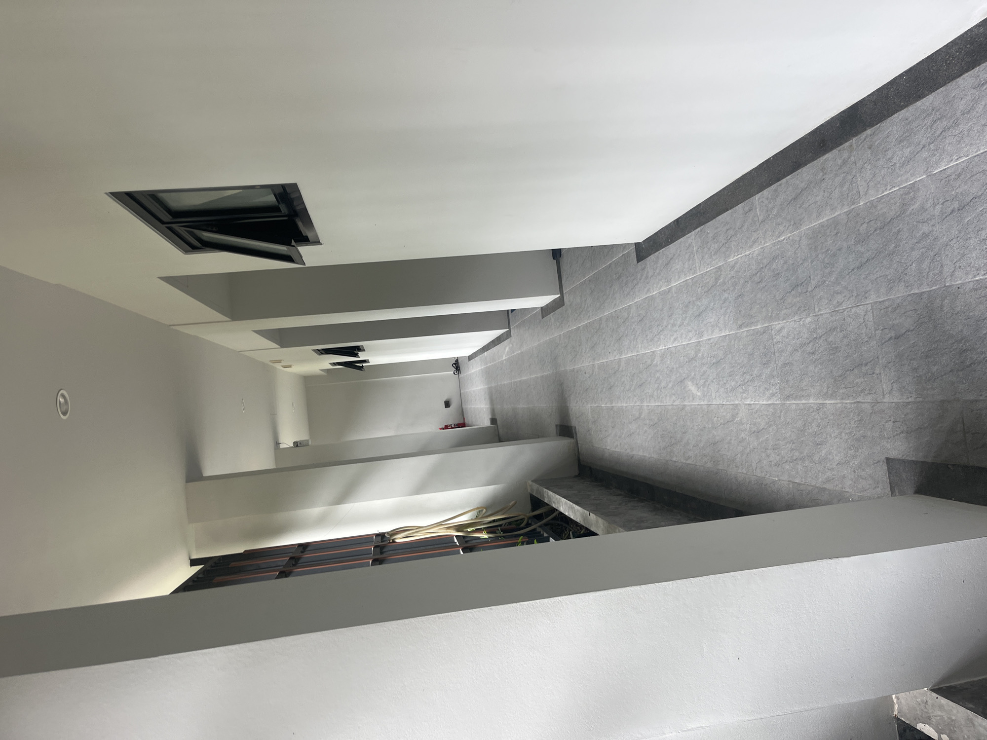 The image shows a clean, modern outdoor hallway in Rawai, Thailand. The hallway is lined with white walls and features gray tiled flooring. On the left side, there is a planter box containing some greenery and a coiled hose. Further down the hallway, two open windows are visible on the right wall, and a red fire extinguisher is mounted on the wall on the left. The hallway extends into the distance, suggesting it is part of a building complex or a row of rooms. The lighting is bright, indicating it is daytime.