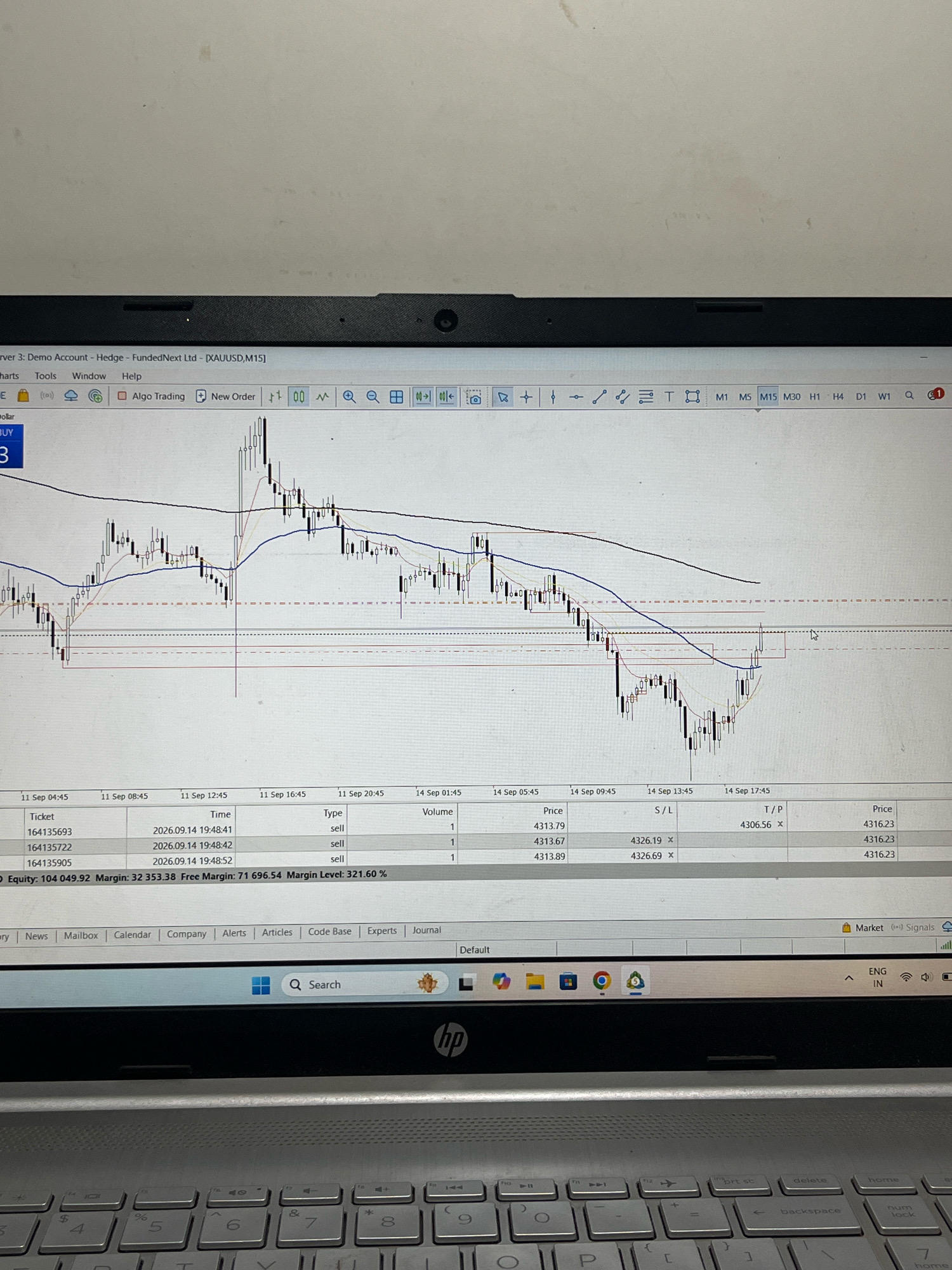 This image shows a laptop screen displaying a trading platform, likely for financial markets. The screen exhibits a candlestick chart with various technical indicators like moving averages, and a table of trade data including ticket numbers, time, type (sell), volume, price, stop loss (S/L), and take profit (T/P). The platform appears to be MetaTrader 5, indicated by the interface elements and the XAUUSD currency pair.
The scene is set indoors, with the laptop placed on a silver-colored keyboard. The background is a plain, light-colored wall. There are no people visible in the image, nor any specific environmental features that would allow for the determination of a city, including Hiangtam Lamka, India. The image focuses solely on the laptop screen and keyboard.