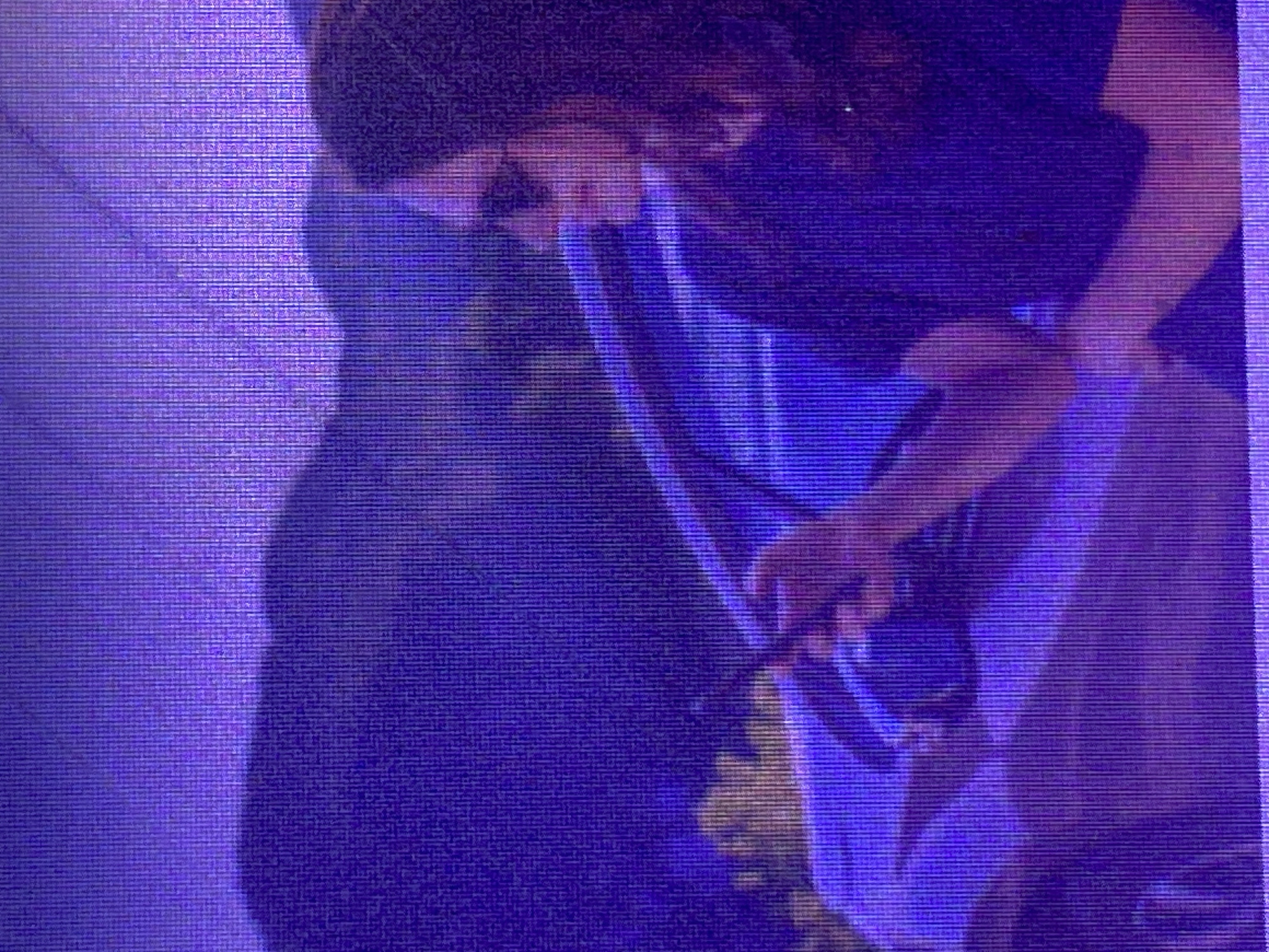 The media file shows a close-up, horizontally framed, dimly lit shot of a person from the chest up, leaning forward. The person is wearing a dark beanie and a dark shirt. Their right hand is extended, holding a dark object that appears to be a camera or a similar electronic device. The background is largely obscured by shadow and purple and blue light, suggesting an indoor setting, possibly a stage or a dimly lit room. The lighting creates a dramatic effect, highlighting the person's face and hands. The image quality is grainy, suggesting it might be a low-resolution photograph or a screenshot from a video. There is no visible text in the image. The overall mood is somewhat mysterious and focused on the act of capturing an image or recording something. The location context of Ikorodu, Nigeria, does not provide specific visual cues within this image.