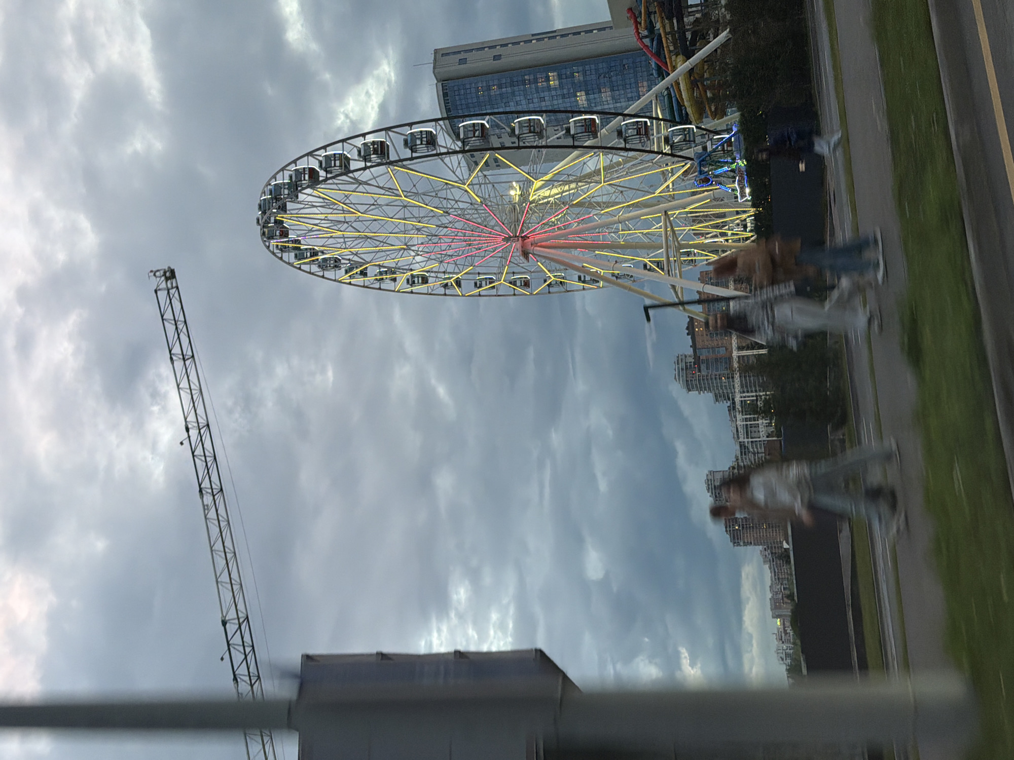 The image shows a Ferris wheel at an amusement park, with a cloudy sky overhead. In the foreground, a construction crane is visible. In the background, there are tall buildings, suggesting an urban setting. The Ferris wheel is illuminated with yellow and pink lights, and several gondolas are visible. A few people are blurred in motion on a path next to the amusement park, suggesting they are walking or moving. The time of day appears to be late afternoon or early evening, given the cloudy conditions and the lighting of the Ferris wheel. The weather is overcast. The scene is likely in an urban area in Kazan, Russia, as indicated by the architecture and the presence of an amusement park. There is no visible text in the image.