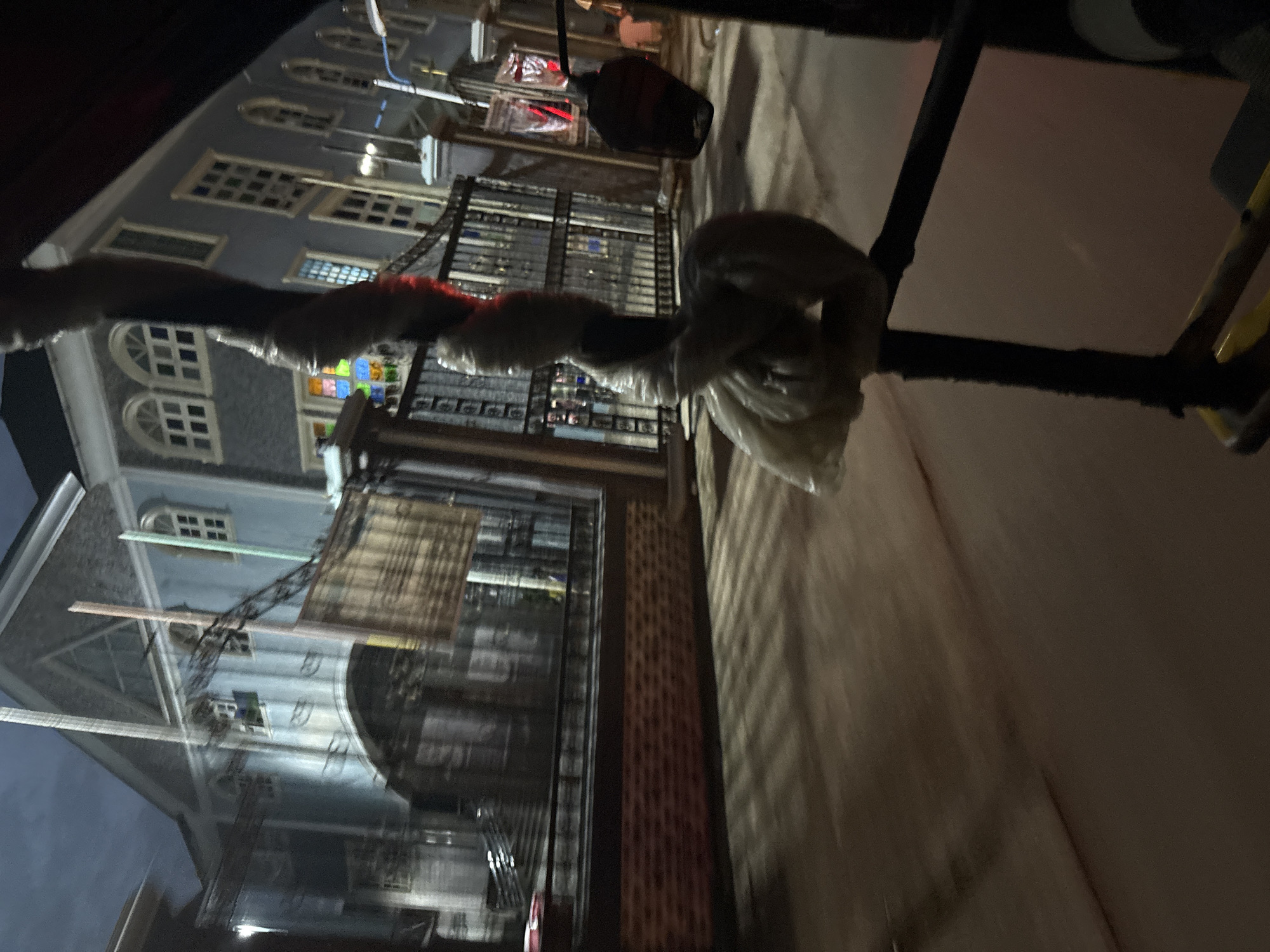 The image depicts a street scene in Onitsha, Nigeria, at dusk or night. The foreground is dominated by a dark, blurry object wrapped in plastic, possibly part of a vehicle or structure. Behind this, a wrought-iron fence runs along a sidewalk or street. Beyond the fence, a light blue building with arched windows and multiple panes is visible. The building appears to be illuminated from within, casting light onto the street. A sign, possibly made of bamboo or wood, hangs from a metal pole in front of the building. The street surface is paved and shows reflections of light, indicating it may be wet or have a glossy finish. In the background, other structures and streetlights can be seen, contributing to the urban nighttime ambiance.