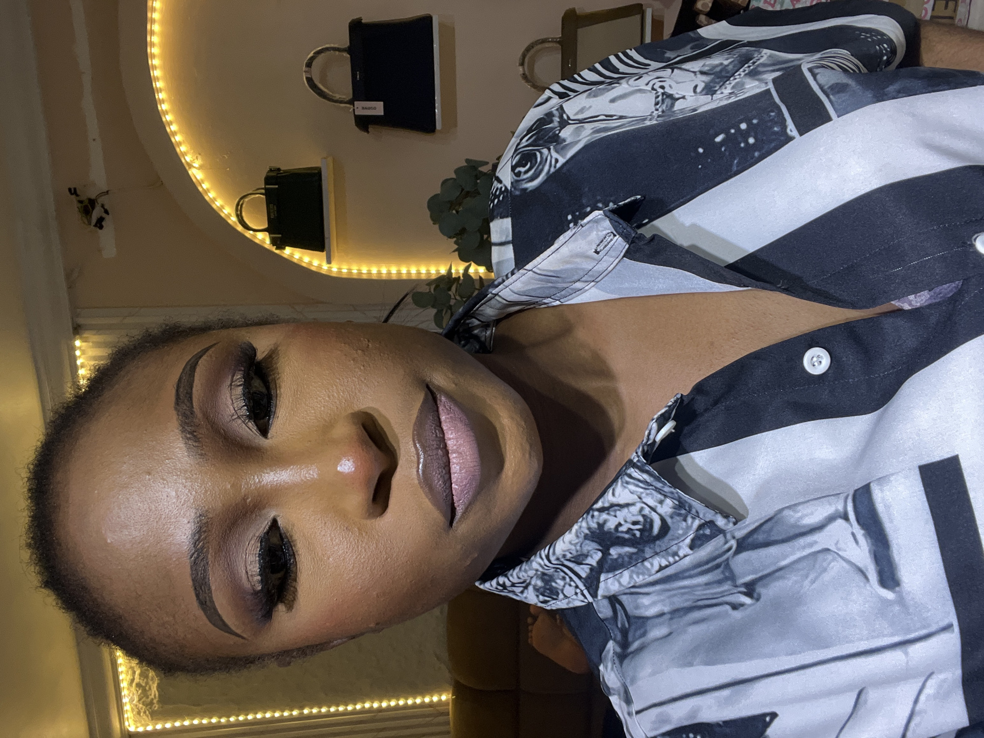 The image is a close-up, selfie-style shot of a woman with dark skin, styled hair, and full makeup. She is wearing a black and white patterned button-up shirt. The background is dimly lit, featuring a shelf with several handbags, and a curved wall illuminated by yellow fairy lights. Behind her to the left, a couch and a hint of feet are visible. The overall impression is that of a person posing for a photograph, likely indoors in a decorated space. The setting appears to be a retail or personal display area, possibly a boutique or a home interior, with the handbags suggesting a fashion focus. There is no information within the image to place the location in Abuja, Nigeria.