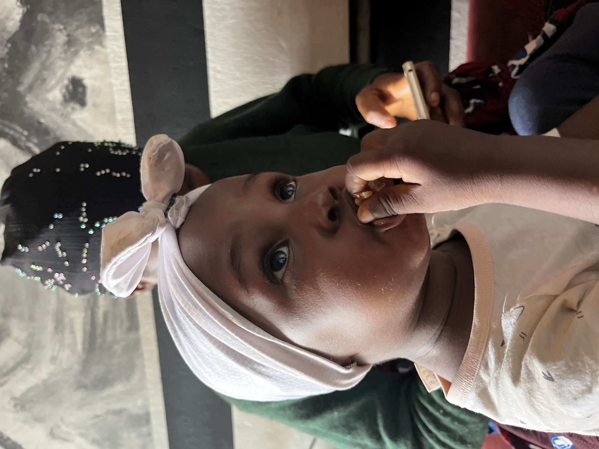 This close-up image, originally rotated 90 degrees clockwise, portrays a young child being fed indoors.
In the foreground, a young child, likely a toddler or preschooler, with dark skin and prominent dark eyes, looks directly towards the camera with a neutral, perhaps curious, expression. The child wears a light-colored head covering, tied into a bow on top, and a light-colored long-sleeved top with subtle dark patterns. A hand, presumably of an unseen feeder, holds small, light-colored pieces of food near the child's open mouth. A tag on the child's collar shows the partial text "LLE".
In the background, the upper body and arm of another individual are visible, wearing a dark green long-sleeved garment. This person is engrossed in a smartphone, whose screen emits a bright glow, illuminating their hand. Behind the child's head, a black fabric adorned with numerous iridescent sequins or beads adds a shimmering detail to the setting. To the far right, a glimpse of red and blue patterned fabric can be seen, possibly part of a cushion or clothing.
The setting appears to be an indoor space, with a wall featuring vertical black and lighter-toned panels or stripes. The lighting suggests an indoor environment, possibly with low natural light or artificial illumination, given the phone screen's glow. The activity centers around the child being fed, while the background figure is occupied with their phone. The image provides no specific location cues other than the general appearance of the subjects, but the provided context places the scene in Barakin Lerawi, Nigeria.