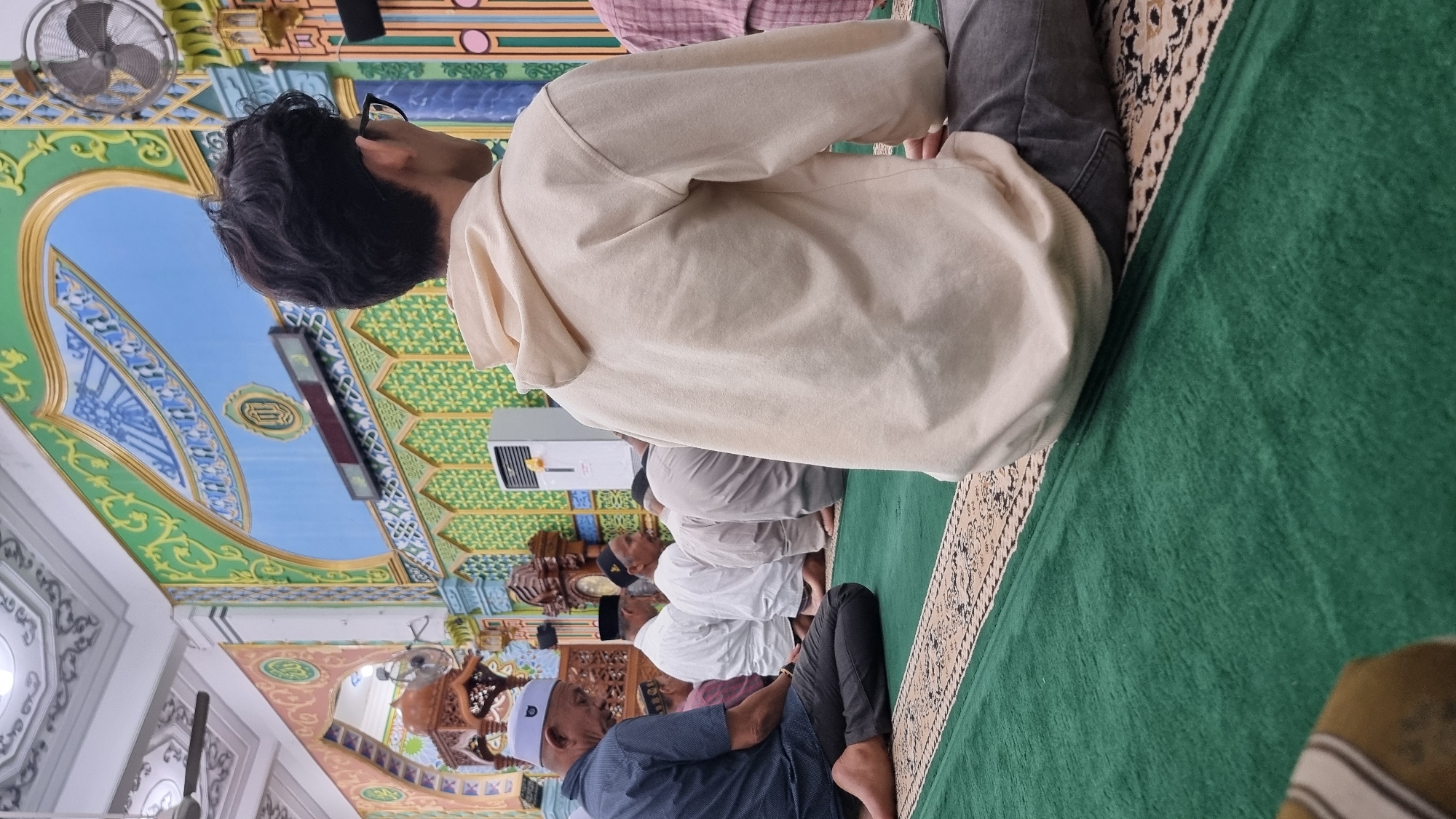 This is a medium shot taken from a low angle inside a mosque in Keudekruenggeukueh, Indonesia, during prayer. In the foreground, a young person with dark, curly hair and glasses is seated on a green carpeted floor, facing away from the camera. They are wearing a light-colored hooded sweatshirt and dark pants. To the left, several men are also seated on the carpet in prayer positions. One man in the left foreground is wearing a dark blue shirt and a white peci cap. The mosque's interior features ornate decorations on the walls and ceiling, with Islamic calligraphy, arches, and patterned designs in shades of green, blue, gold, and pink. A digital clock or display is visible above a patterned wall section. Air conditioning units are mounted on the wall. The overall scene depicts a communal prayer gathering within a richly decorated religious space.