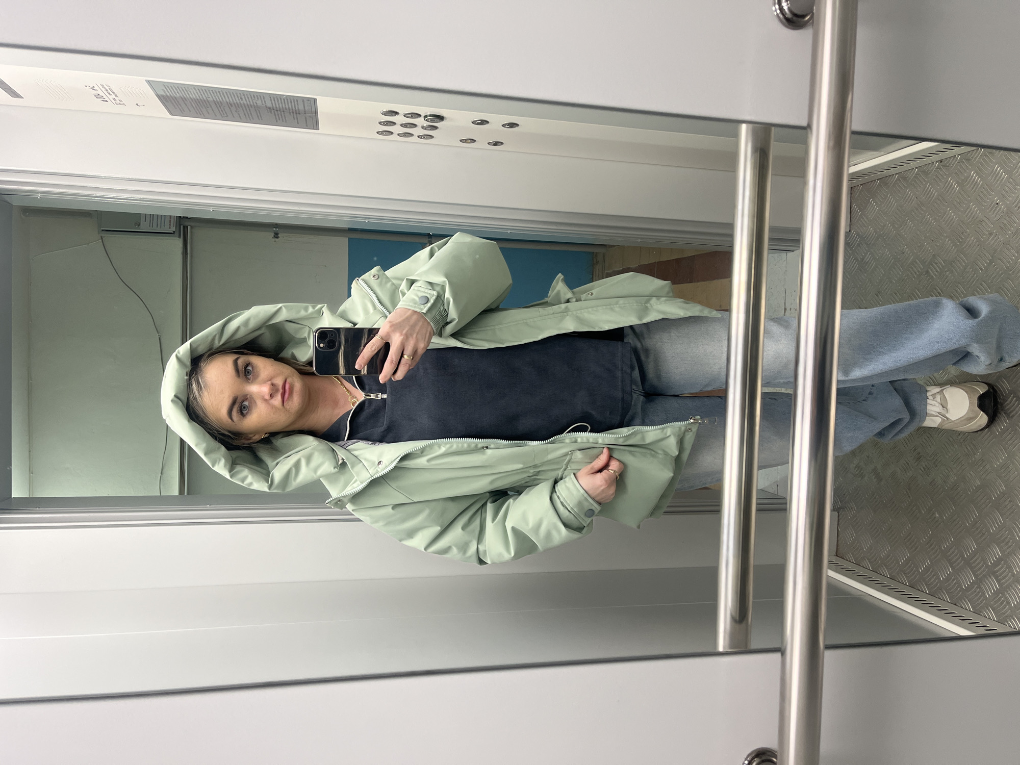 An adult female stands inside an elevator in Chaikovsky, Russia, taking a mirror selfie with a dark-colored smartphone held in her right hand. She wears a light mint green hooded jacket, a dark blue ribbed shirt with a zippered collar, light blue wide-leg jeans, and light-colored sneakers. A gold chain necklace is visible. The elevator features light grey wall panels, a metallic diamond-plate floor, and two horizontal stainless steel handrails. To the right, an elevator control panel with multiple circular buttons and Cyrillic text is visible. The reflection in the mirror shows a hallway beyond the elevator door, characterized by a light blue wall.