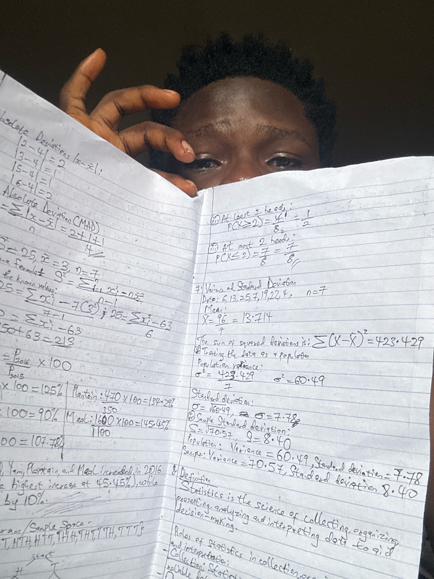 The image displays a close-up view of a person's face and hands holding a notebook filled with handwritten mathematical equations and text. The notebook appears to be a study or work in progress, with various calculations related to statistics. The person's eyes are visible, peering over the top of the notebook, and their hand is partially obscuring the written content. The background is dark and out of focus, suggesting an indoor setting with low lighting. The overall impression is one of study or concentration. The location in Ihiagwa, Nigeria is not evident from the image itself.